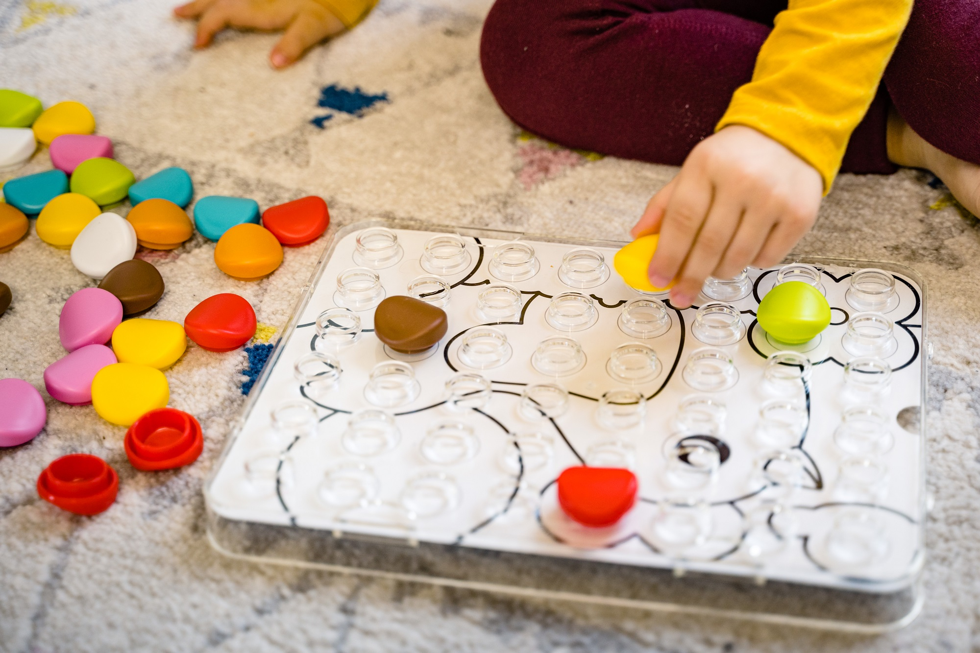 Ein Kind spielt mit bunten Spielzeugteilen auf einem Spielbrett. Die Hand des Kindes platziert ein gelbes Teil.