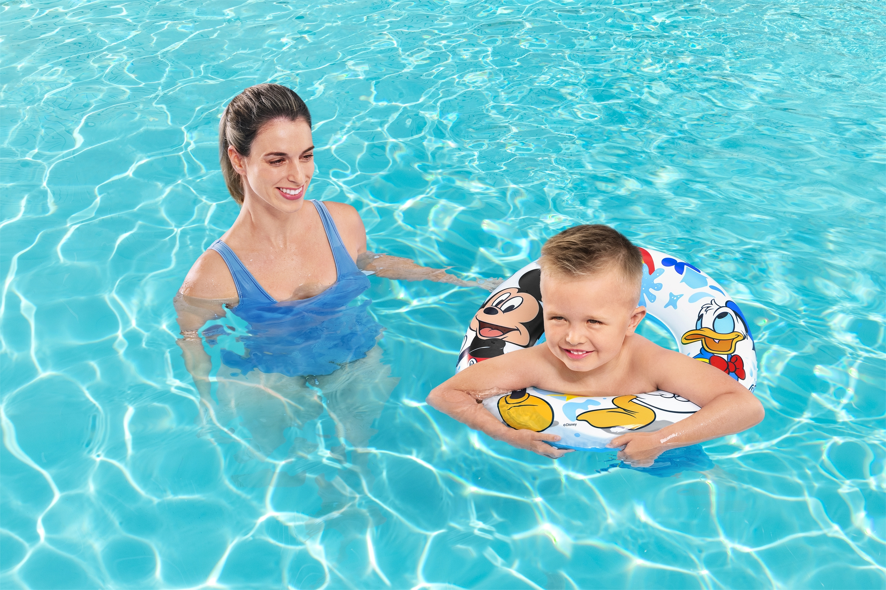 Una mujer y un niño están en una piscina. El niño está en un flotador de Mickey Mouse. El agua es azul.