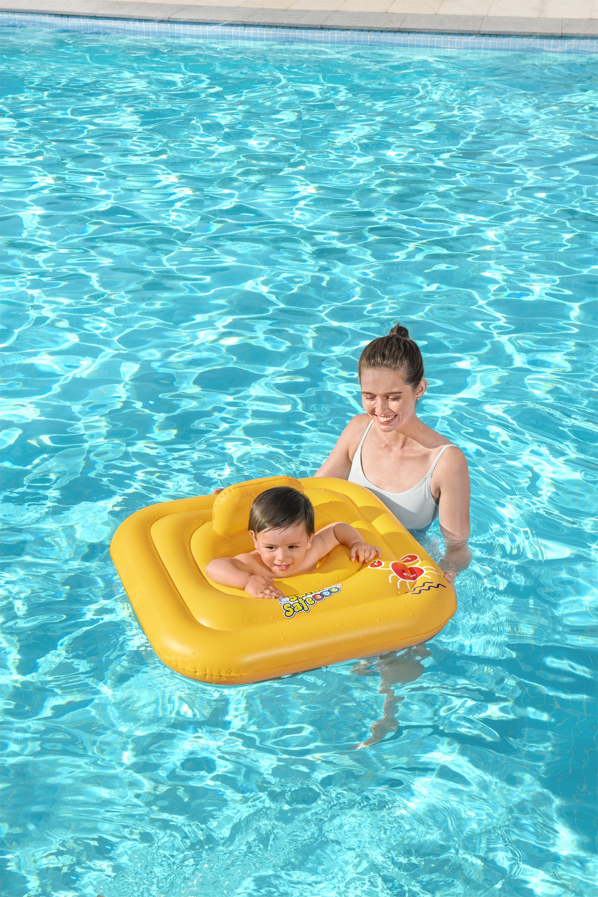 Eine Frau und ein Baby sind in einem Pool. Das Baby sitzt in einem gelben aufblasbaren Ring. Das Wasser ist blau.