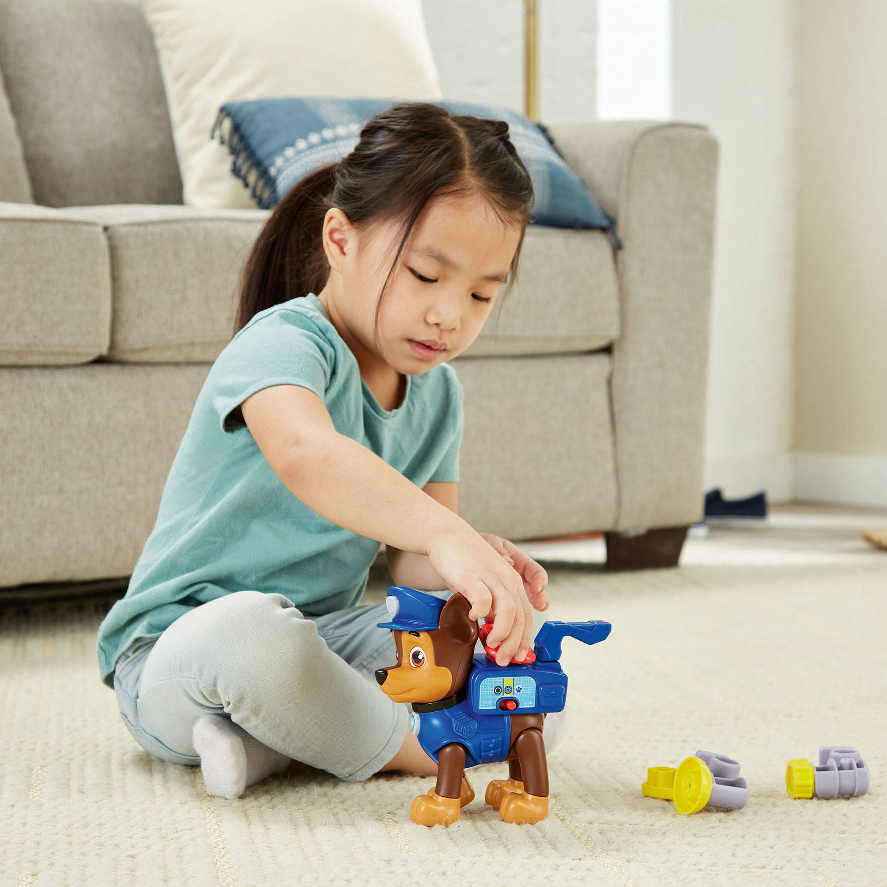 Una bambina gioca con un giocattolo di Chase, seduta su un tappeto. Il giocattolo è un personaggio di Paw Patrol. Un divano è sullo sfondo.