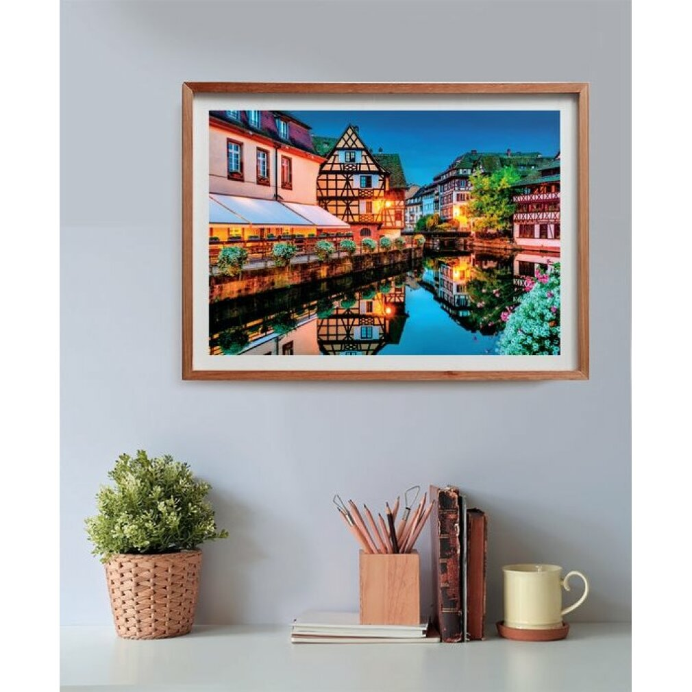 Een ingelijste afbeelding van een Europese stad met kanalen. Gebouwen weerspiegelen in het water. Een plant en bureau-artikelen op de voorgrond.