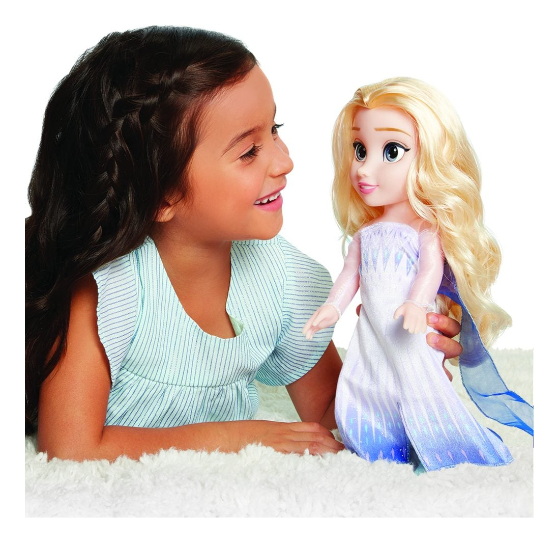 Una niña sonríe a una muñeca Elsa. La muñeca se sostiene sobre una superficie blanca y esponjosa. La niña lleva una camisa a rayas azules.