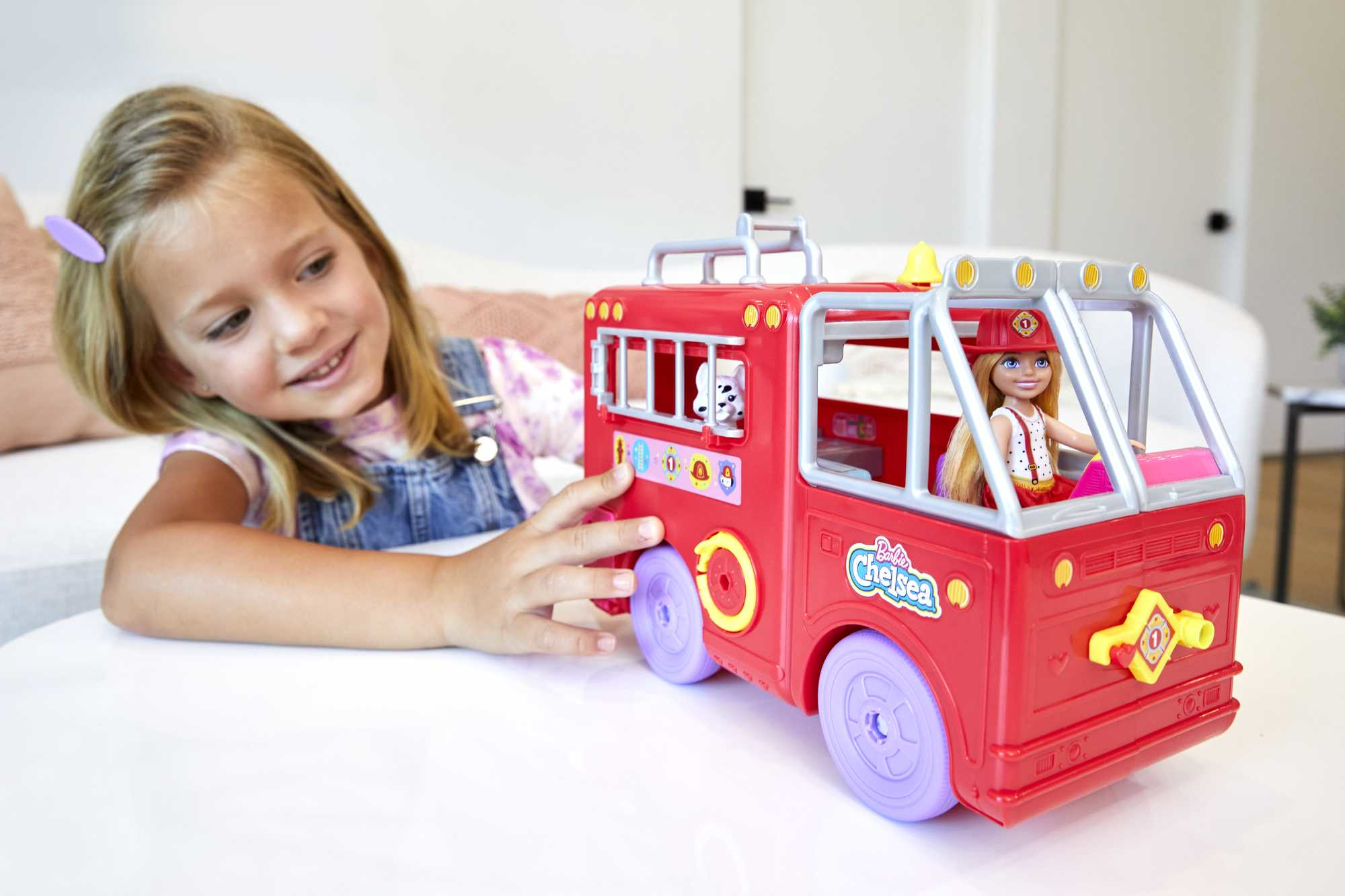 Una bambina sorride a un camion dei pompieri giocattolo rosso. Una bambola con un cappello da pompiere è dentro. Un giocattolo dalmata sbuca.