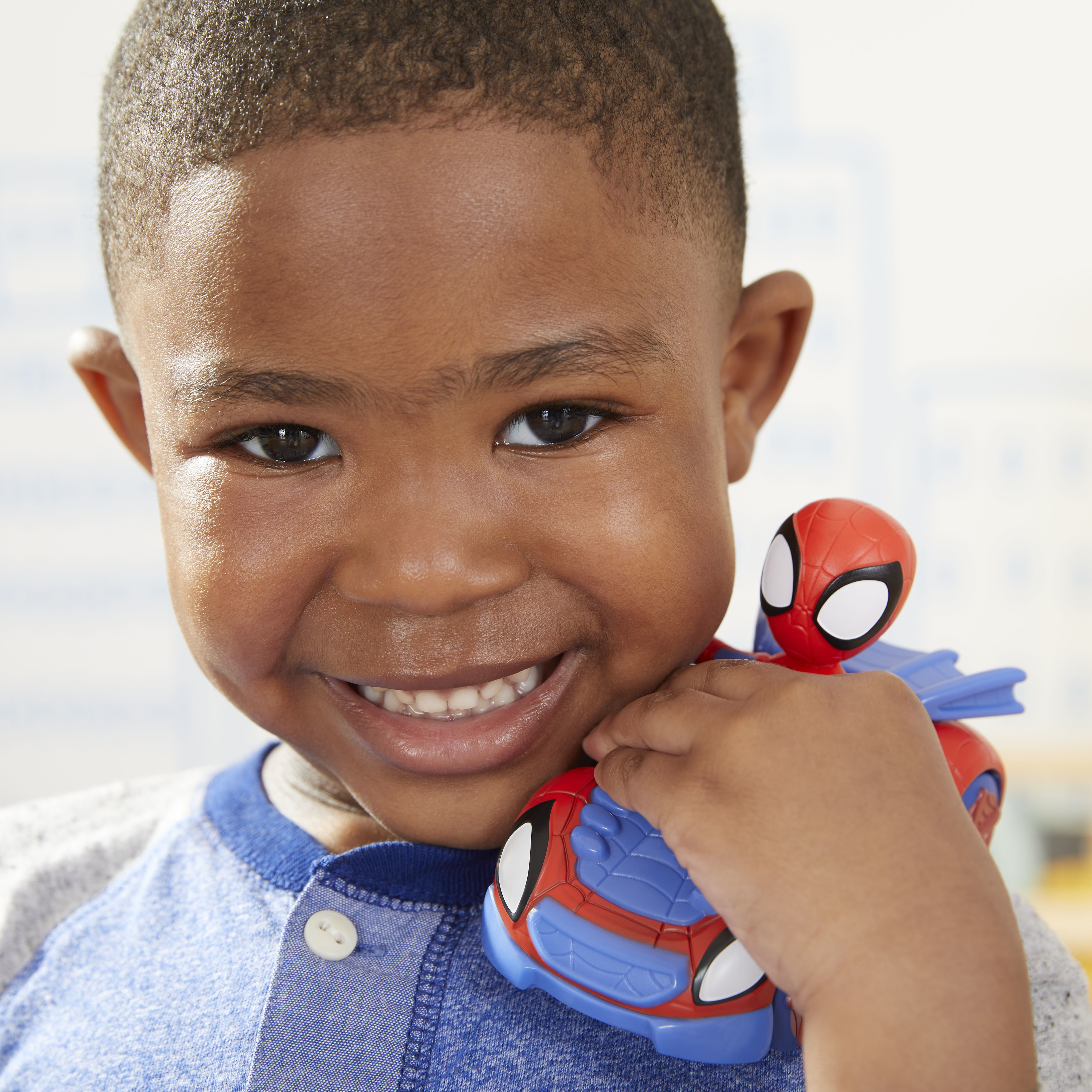 Un niño sonriente sostiene un coche de juguete de Spider-Man. El coche es rojo y azul. El niño lleva una camisa azul.