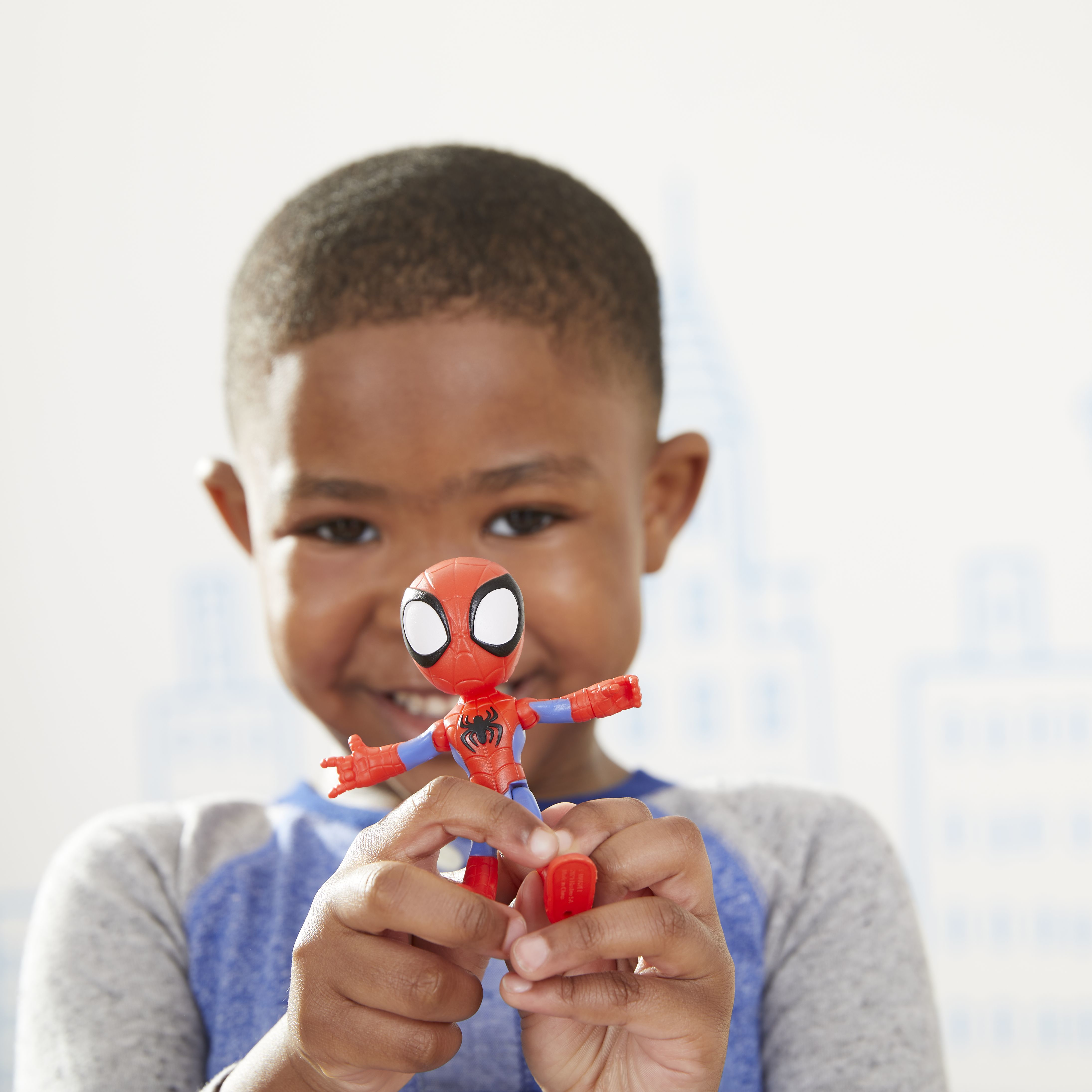 Un niño sostiene un juguete de Spider-Man. El juguete es rojo y azul, y el niño sonríe. El fondo es azul claro con contornos de ciudad.