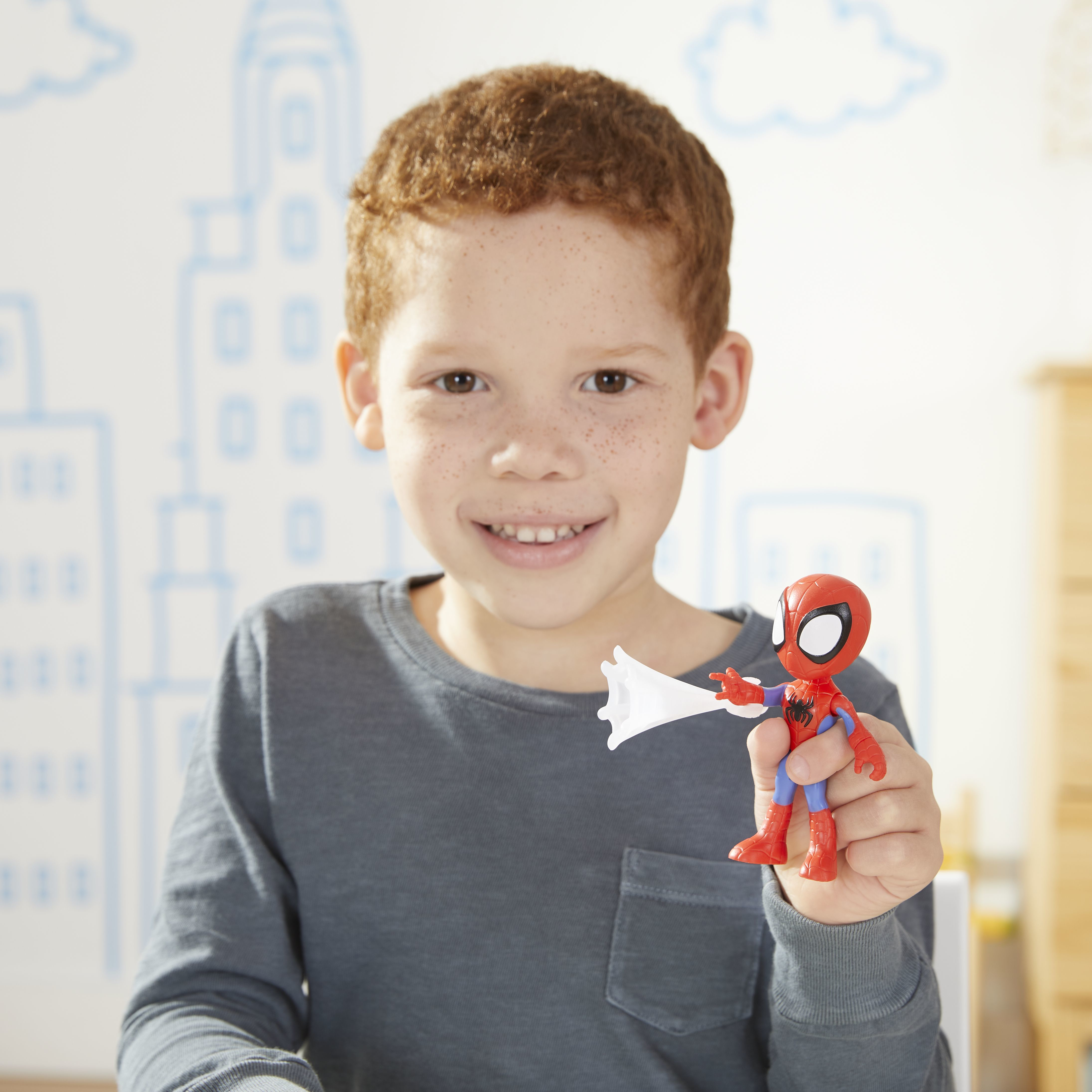 Un niño con cabello rojo sostiene un juguete de Spider-Man. Sonríe a la cámara. Una ciudad está dibujada en la pared.