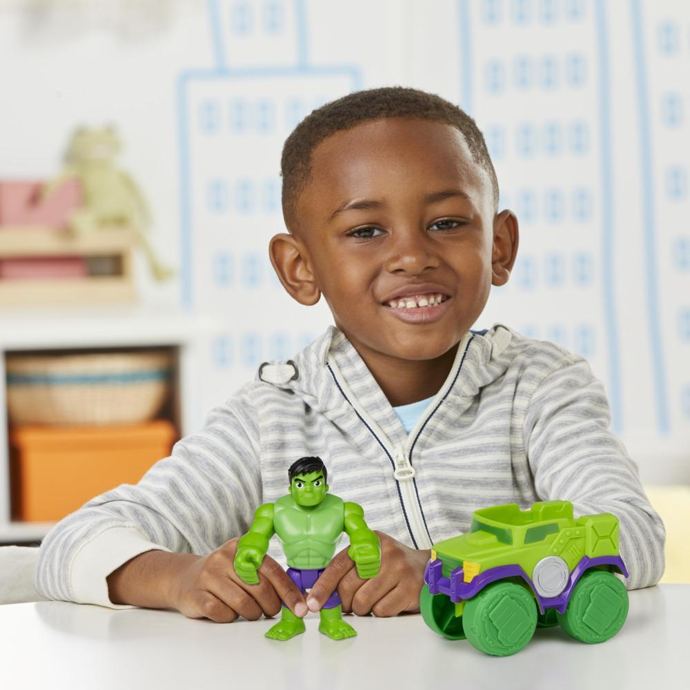 Ein lächelnder Junge mit einem Spielzeug-Hulk und einem grünen Auto. Der Hulk ist grün und lila. Das Auto ist grün, lila und gelb.