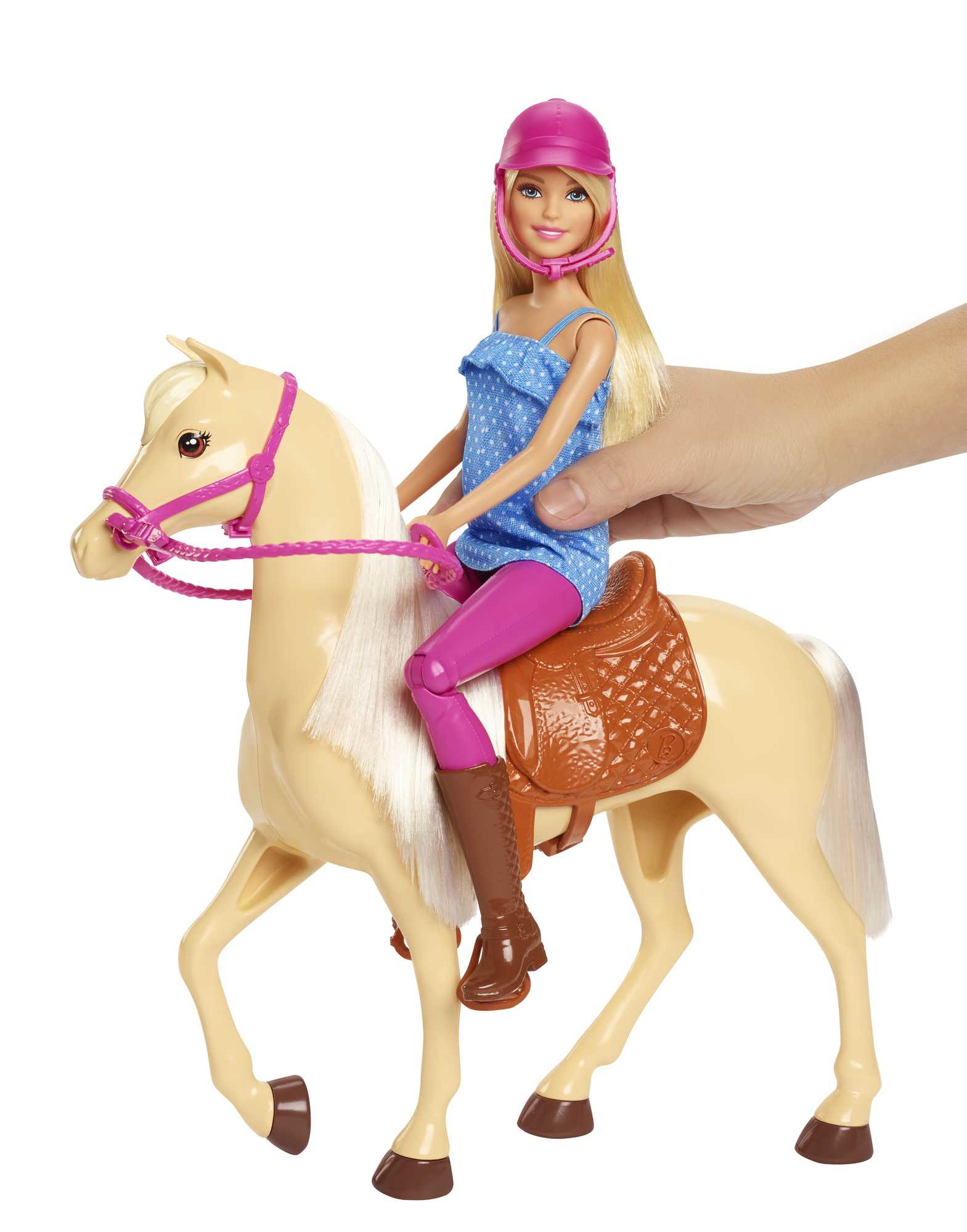 Barbie-Puppe mit rosa Helm reitet auf einem beigen Spielzeugpferd. Eine Hand hält das Pferd. Die Puppe trägt ein blaues Oberteil und pinke Hose.