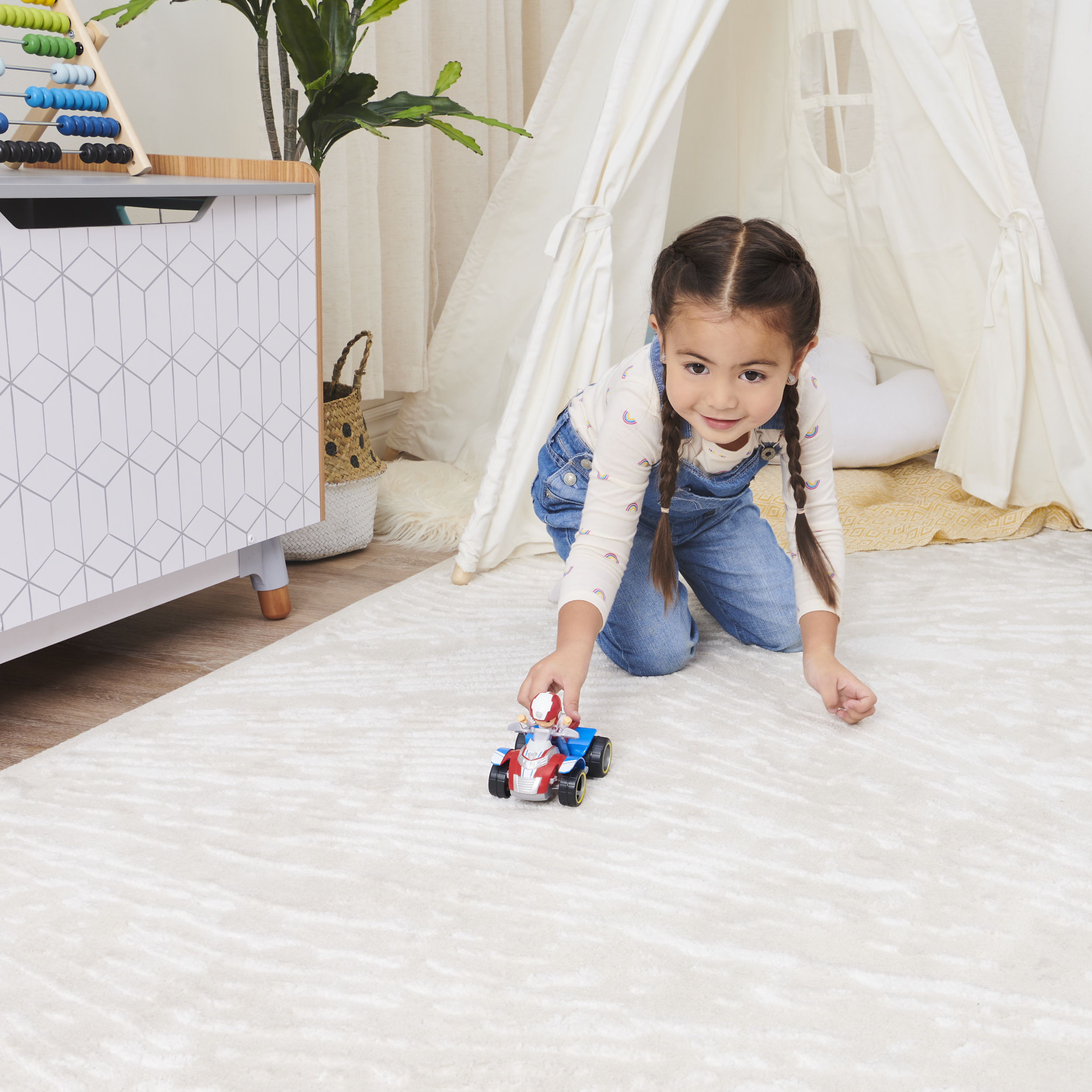 Una niña juega con un coche de juguete sobre una alfombra. Una tienda tipi está en el fondo.