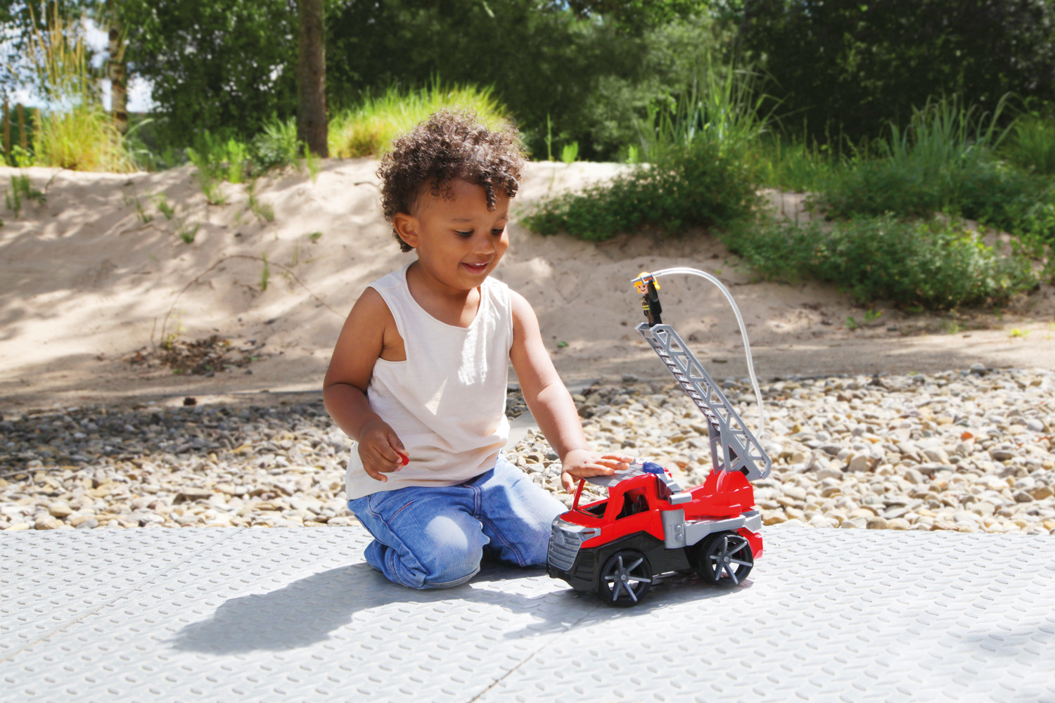 Dziecko klęczy obok czerwonej zabawki wozu strażackiego. Dziecko ma na sobie dżinsy i białą koszulę. W tle piasek i drzewa.
