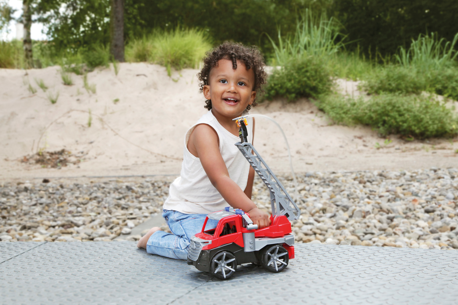 Uśmiechnięte dziecko bawi się czerwoną i szarą zabawką wóz strażacki na teksturowanej powierzchni. Tło obejmuje drzewa i piasek.