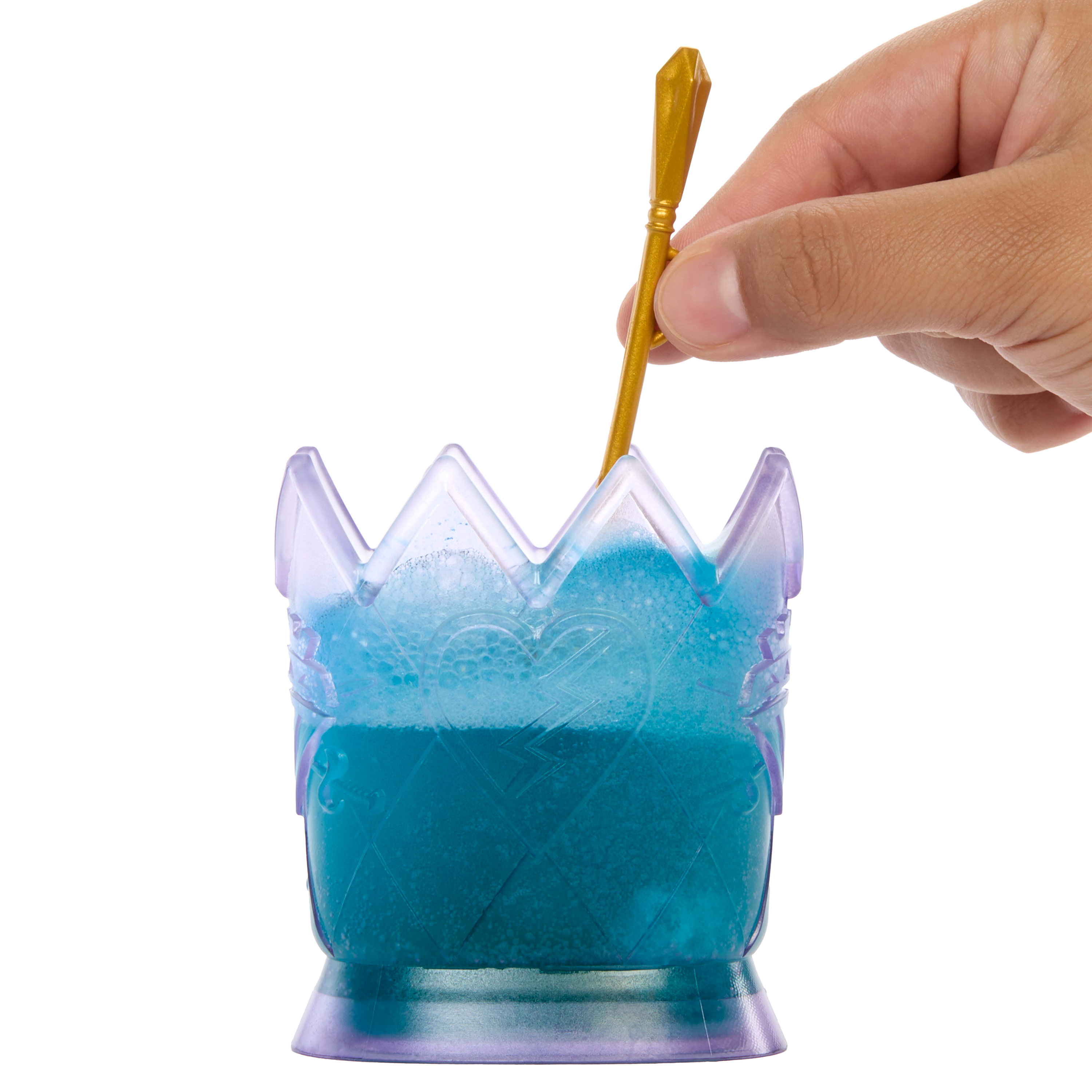 Dłoń miesza złotą łyżką w niebieskiej cieczy w pojemniku w kształcie korony, na białym tle.