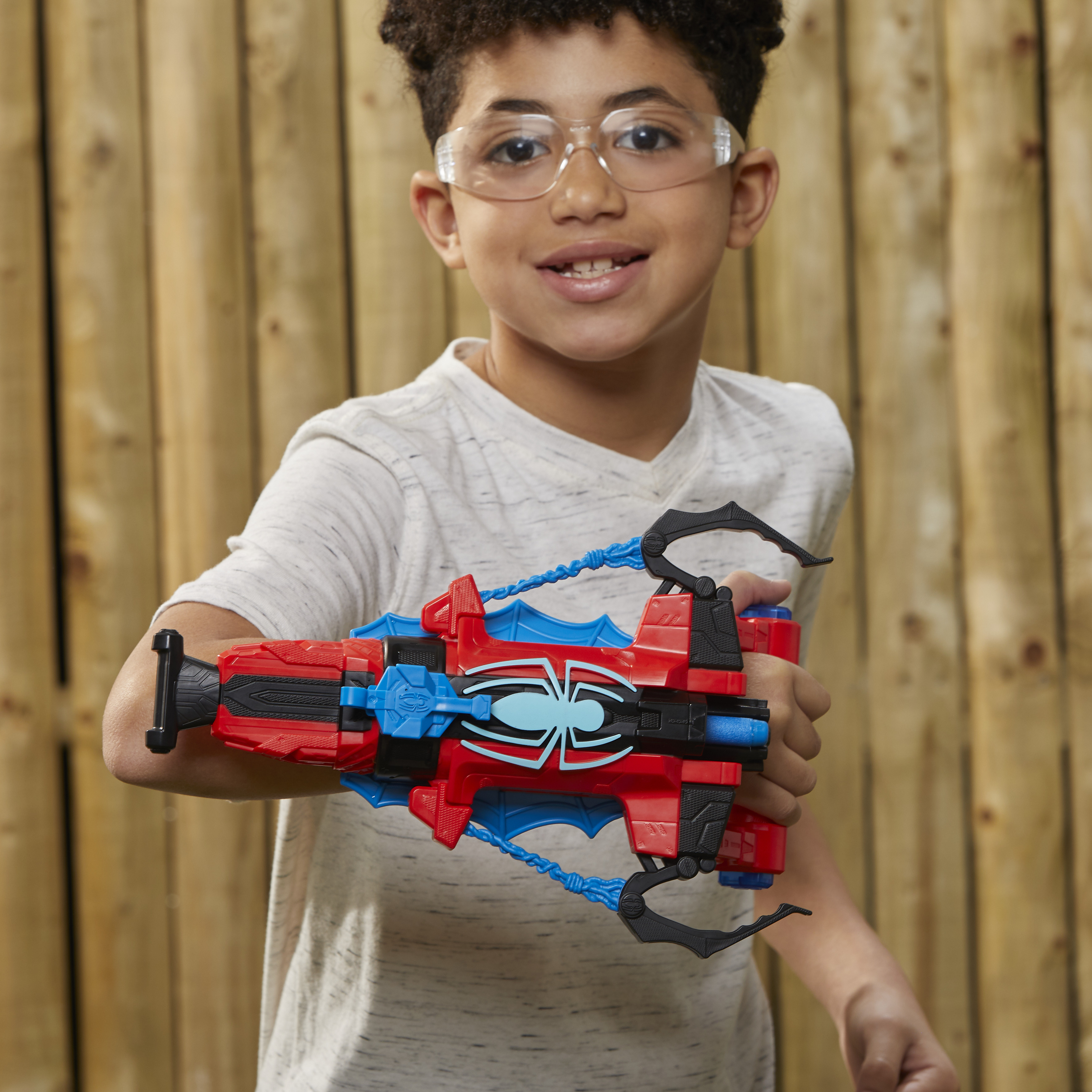 Un niño con gafas sostiene una pistola de juguete de Spider-Man roja y azul frente a un fondo de madera, sonriendo.