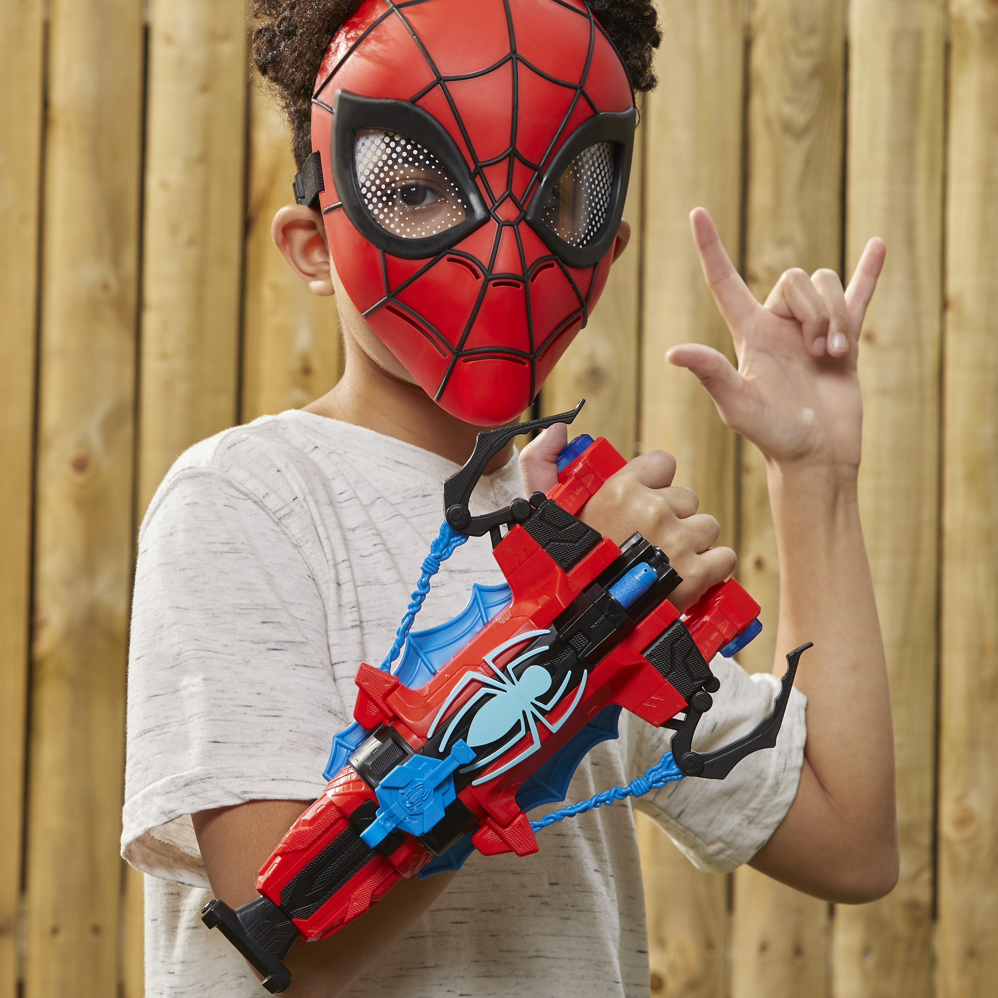 Un niño con máscara de Spider-Man y pistola de juguete, posando frente a una valla de madera.