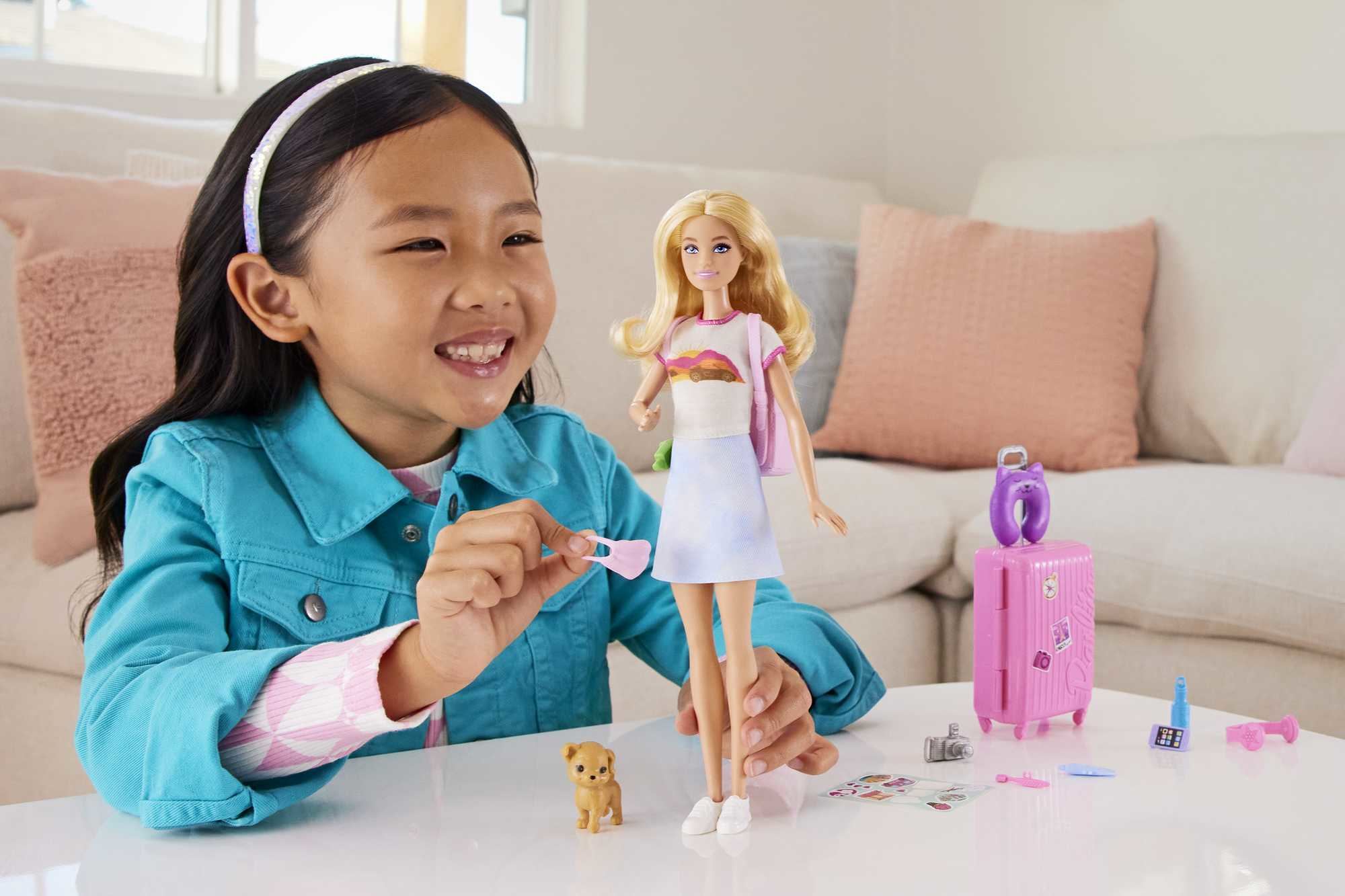 Una niña sonríe a una muñeca Barbie con una bolsa rosa. Los accesorios incluyen una maleta, un perro de juguete y otros objetos pequeños.