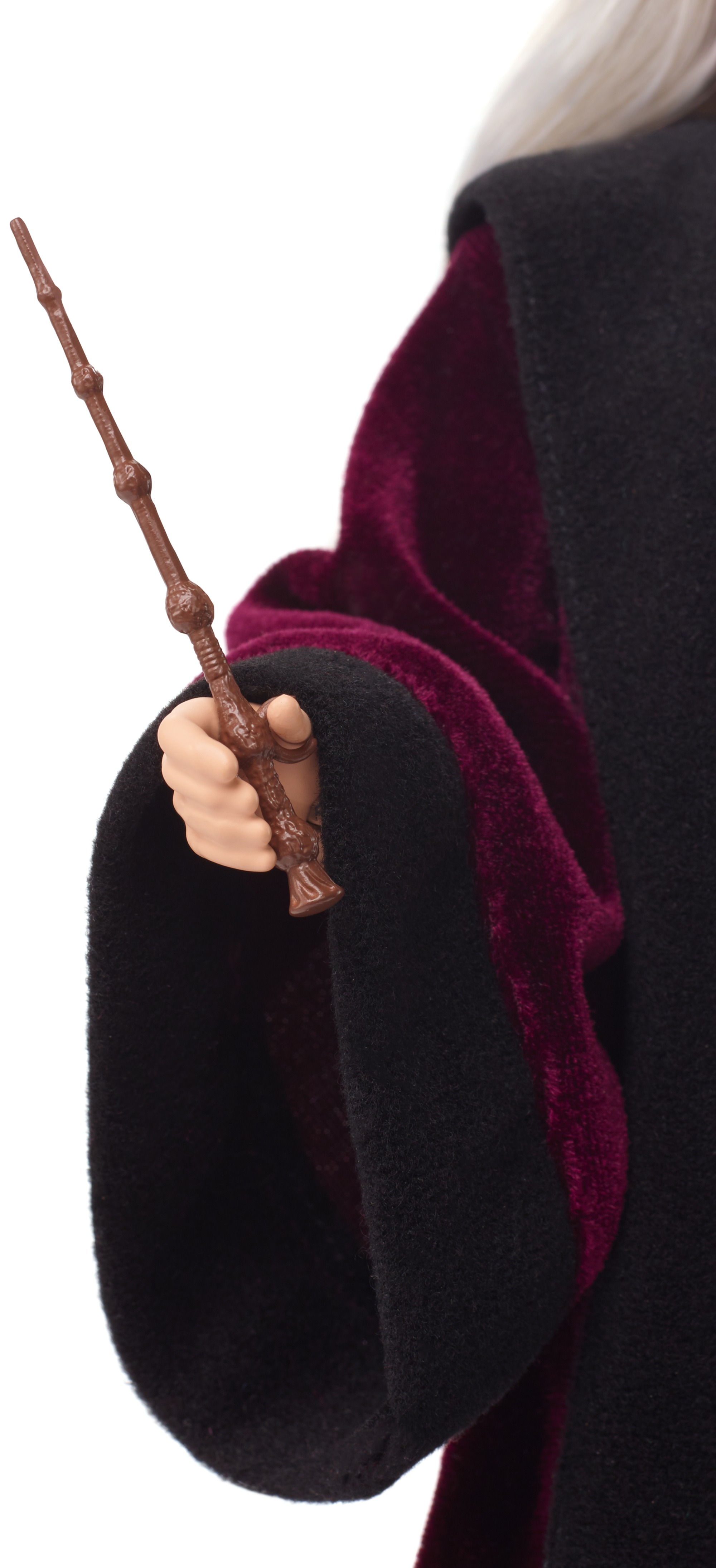 Una mano tiene una bacchetta marrone, parte di una figura in abito nero con fodera in velluto bordeaux. Sfondo bianco.