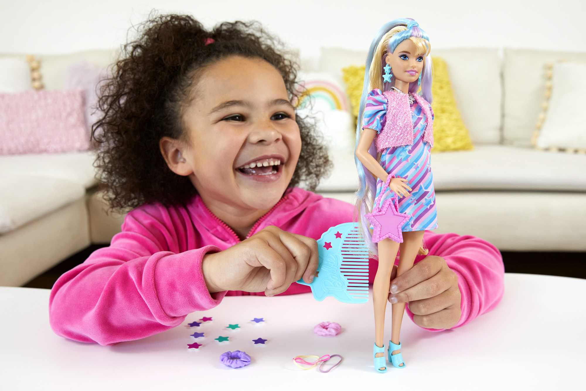 Ein Mädchen lächelt eine Barbie-Puppe an. Die Puppe hat blaue Haare und ein gestreiftes Kleid. Konfetti und Accessoires liegen auf dem Tisch.