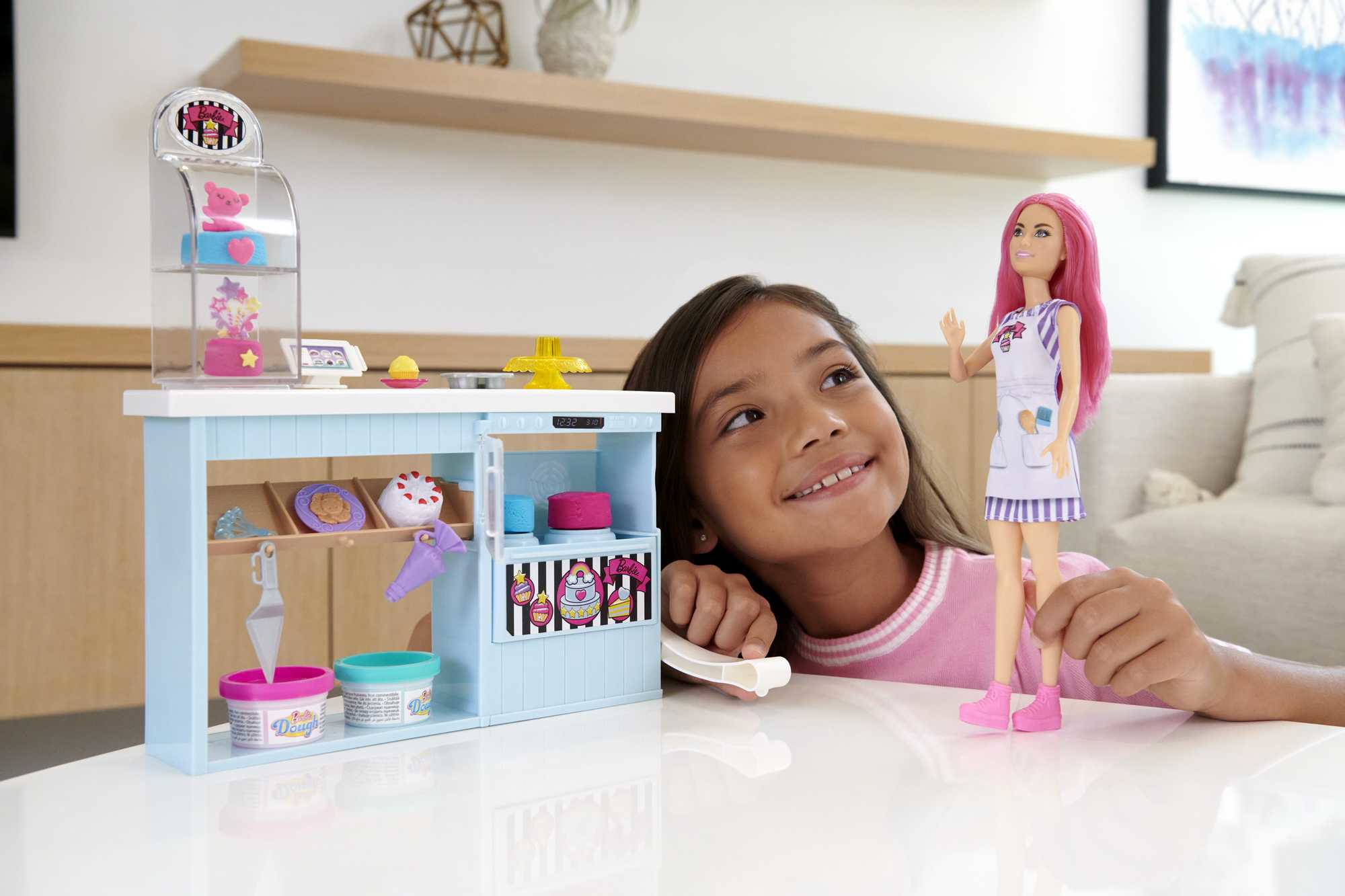 Ein Mädchen lächelt eine Barbie-Puppe mit rosa Haaren an, die neben einem Spielzeugbäckerei-Set mit Gebäck steht.