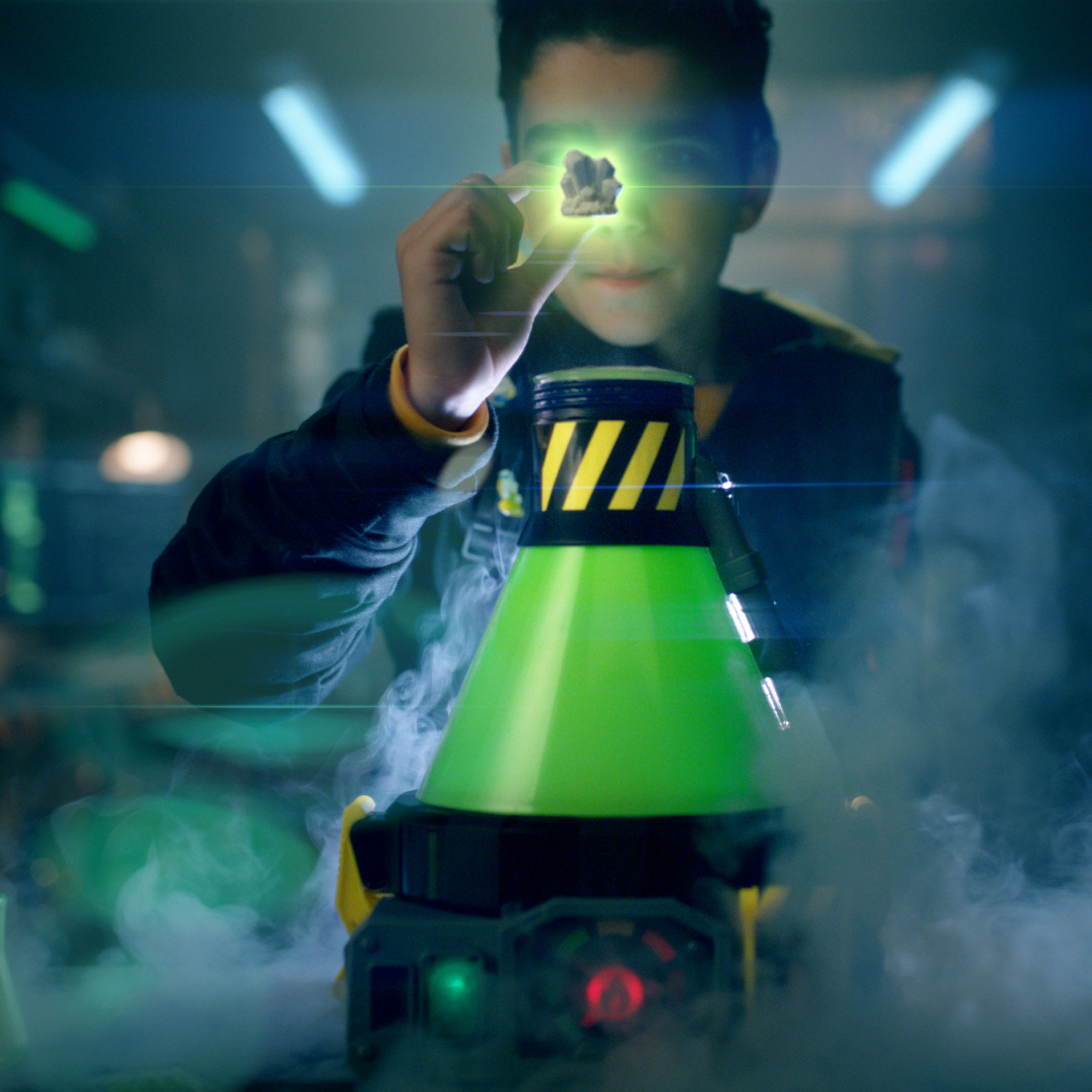 Chłopiec trzyma świecącą skałę. Z przodu zielone, czarne i żółte urządzenie. Obecny dym i niebieskie światła.