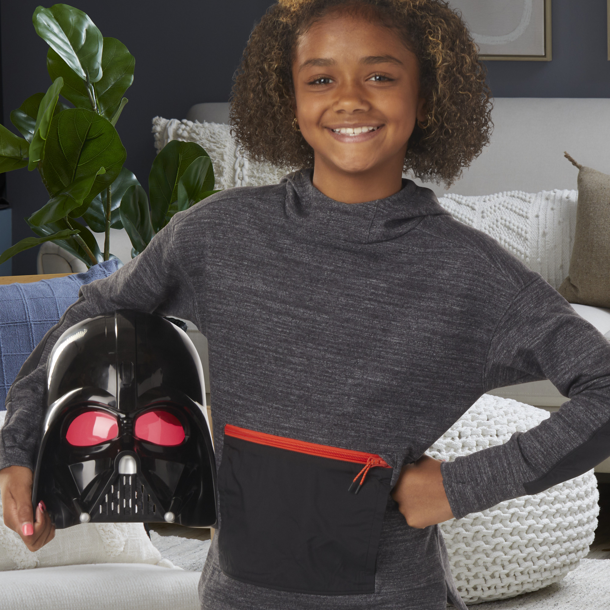 Una niña sostiene una máscara de Darth Vader. Sonríe, lleva una sudadera gris, frente a un salón.