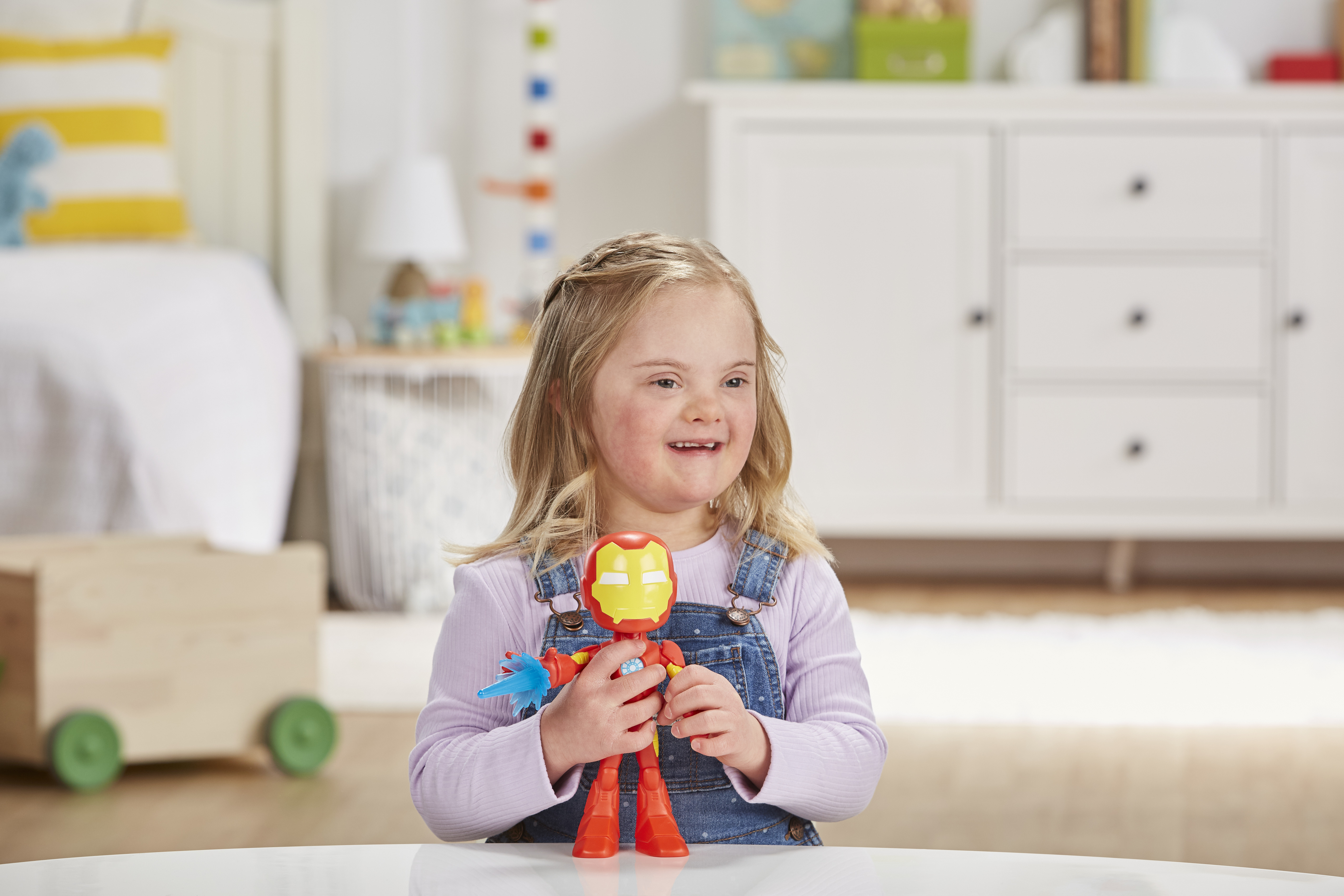 Una bambina con sindrome di Down tiene in mano un giocattolo di Iron Man. Indossa una tuta di jeans e sorride. Un letto e un comò sullo sfondo.