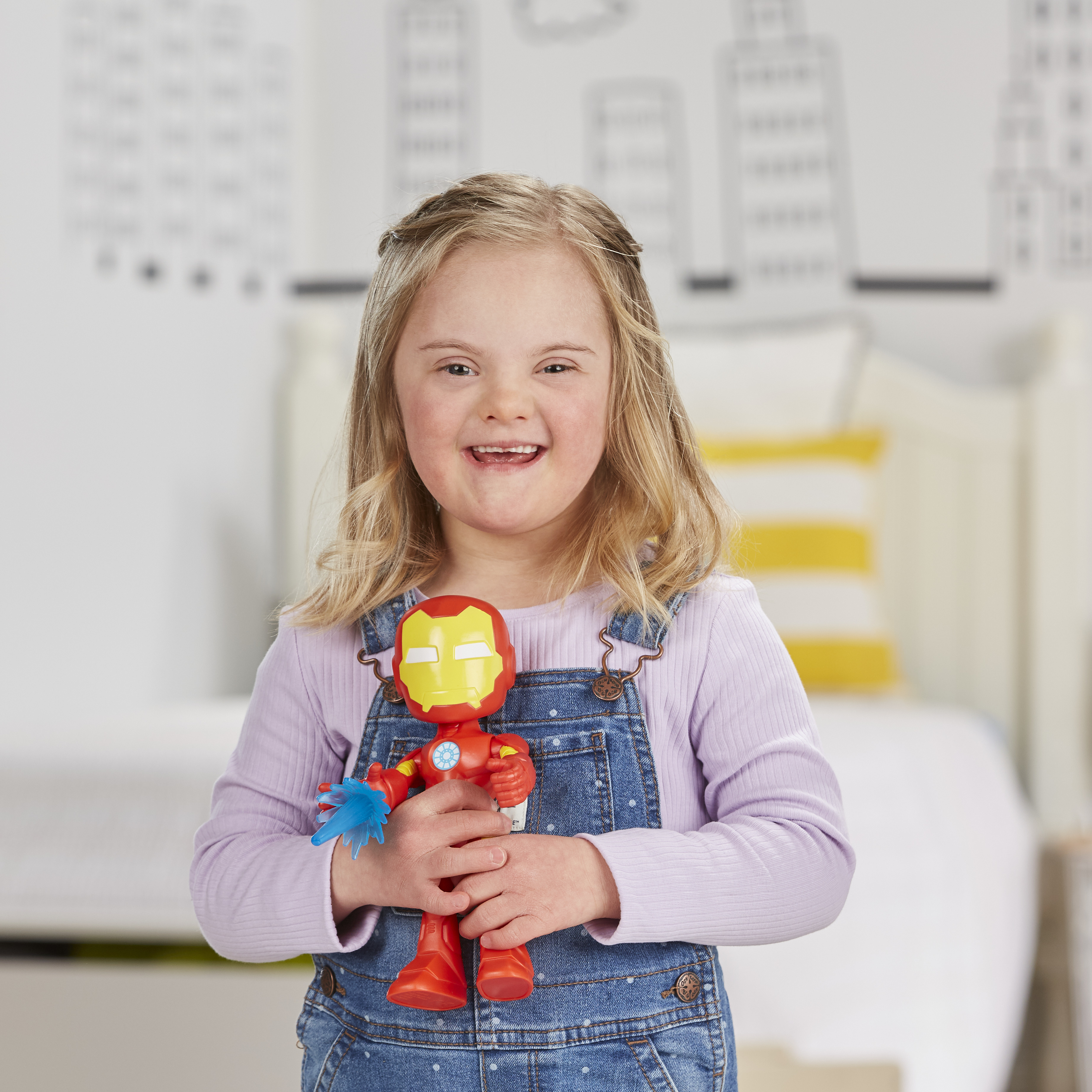 Una bambina sorridente con la sindrome di Down tiene un giocattolo di Iron Man. Indossa una salopette di jeans e una camicia lilla. Sfondo sfocato.