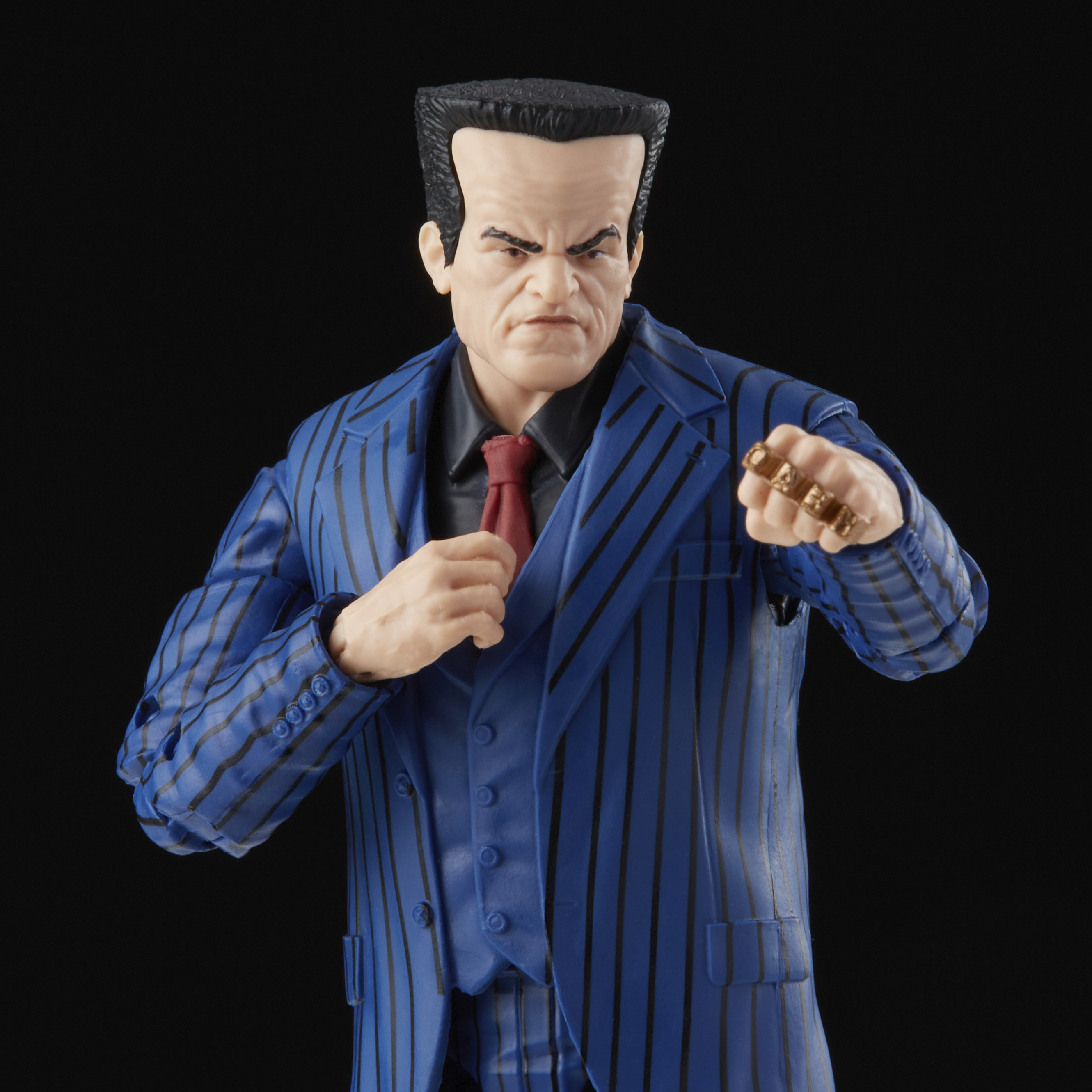 Figura in abito blu gessato, camicia nera e cravatta rossa. Anelli d'oro sul pugno. Sfondo nero.