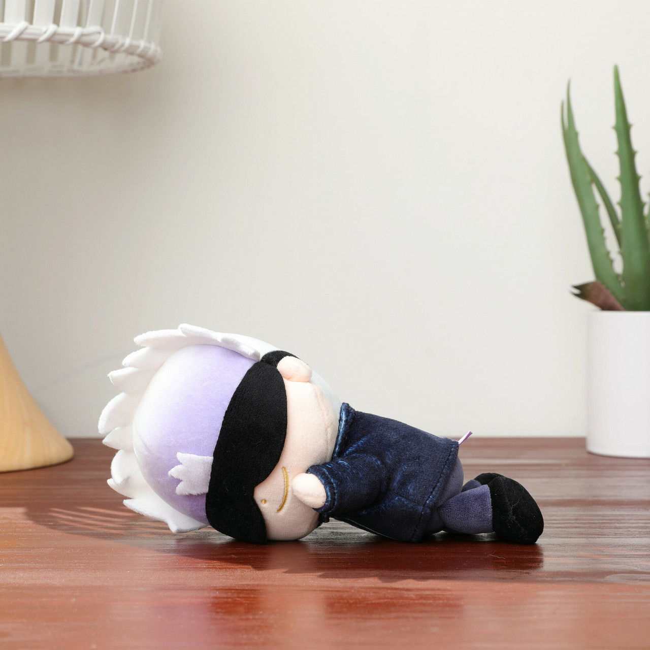Pluszowa zabawka postaci z białymi włosami, czarną przepaską na oczy i ciemnoniebieskim stroju leży na drewnianej powierzchni.
