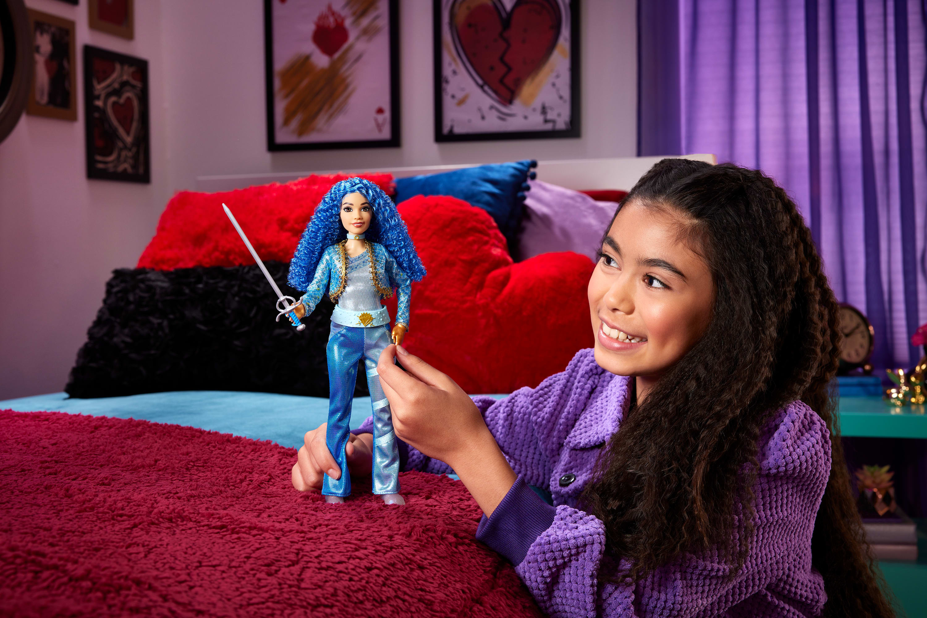 Dziewczyna trzyma lalkę z mieczem. Lalka ma niebieskie włosy. Dziewczyna ma na sobie fioletowy sweter. W tle łóżko.