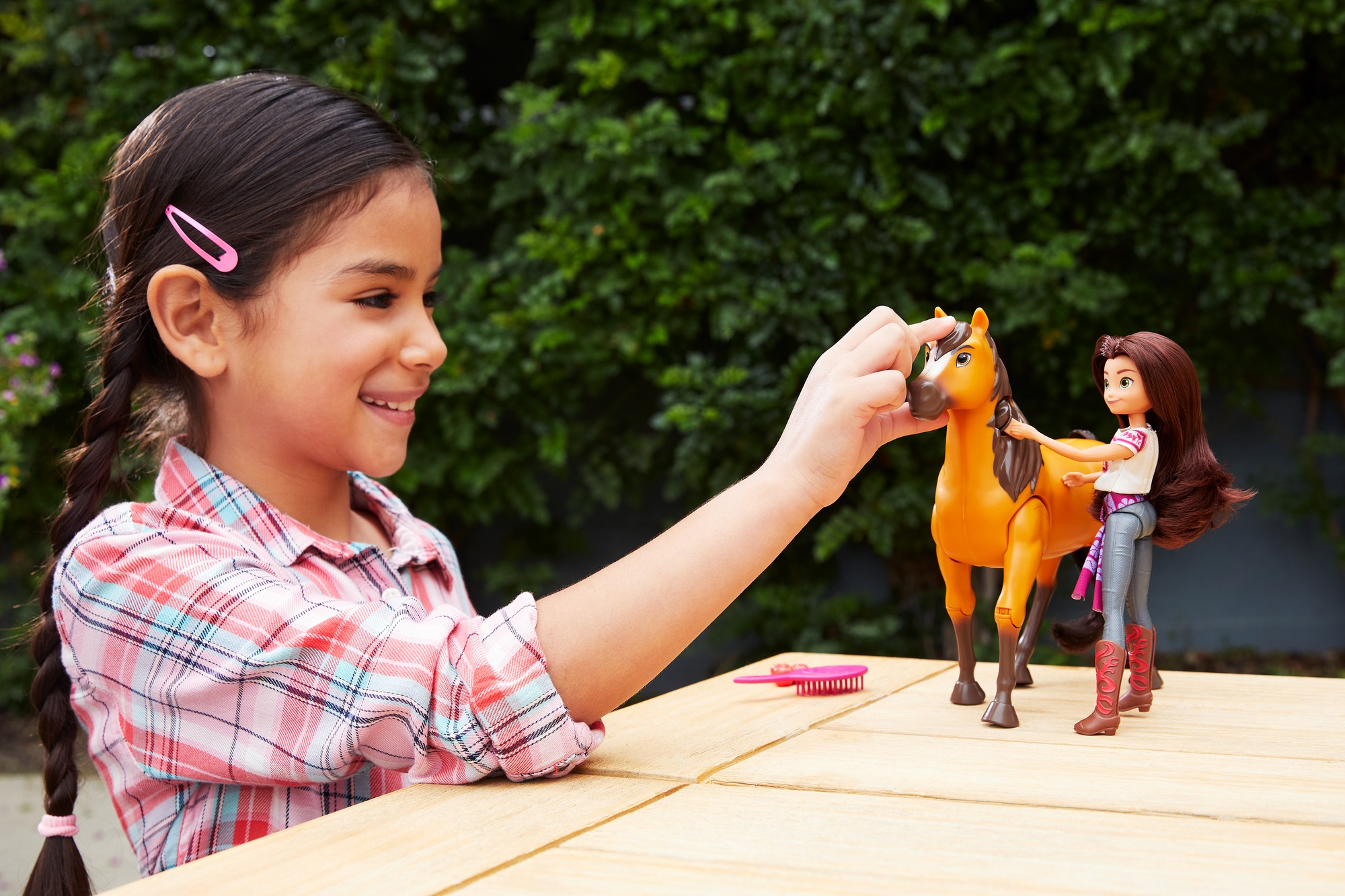 Dziewczyna uśmiecha się, dotykając zabawkowego konia. Lalka stoi obok na drewnianym stole. W tle zielone liście.