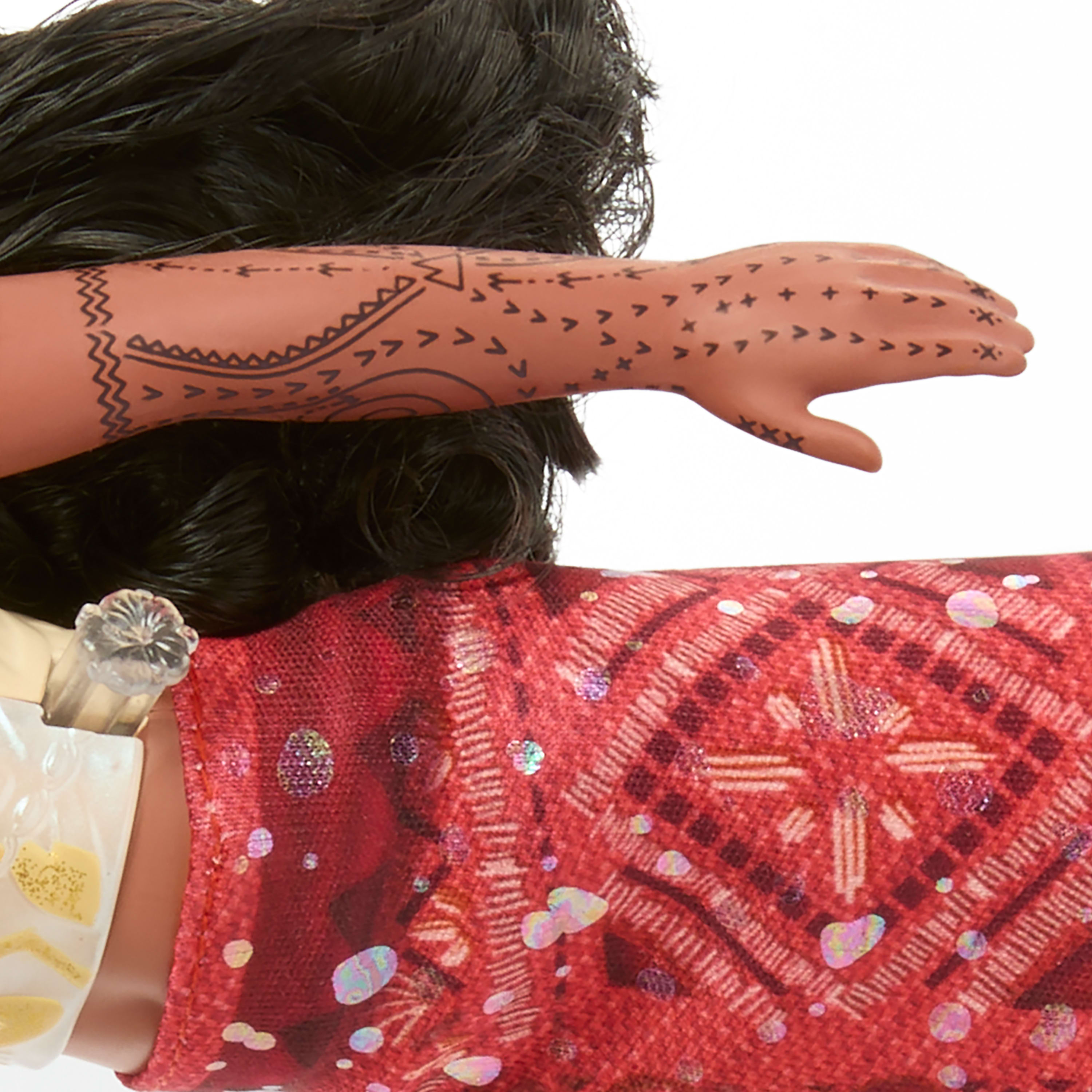 Ramię lalki z tatuażami, czarne włosy i czerwona wzorzysta sukienka. Przezroczysty plastikowy dodatek po lewej.