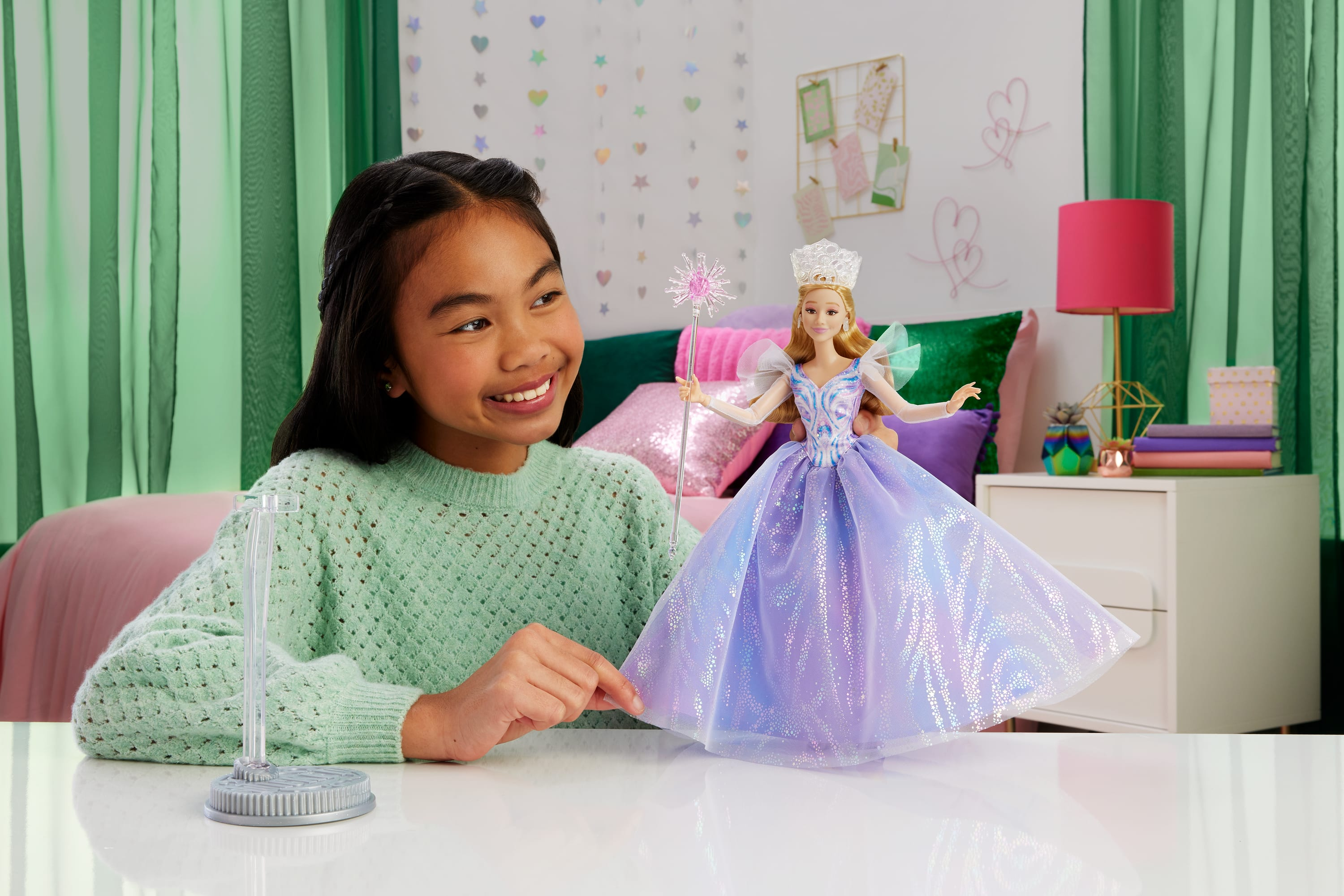 Ein Mädchen lächelt eine Barbie-Puppe in einem lila Kleid an. Dahinter sind ein Bett, Vorhänge und ein Beistelltisch mit Lampe.