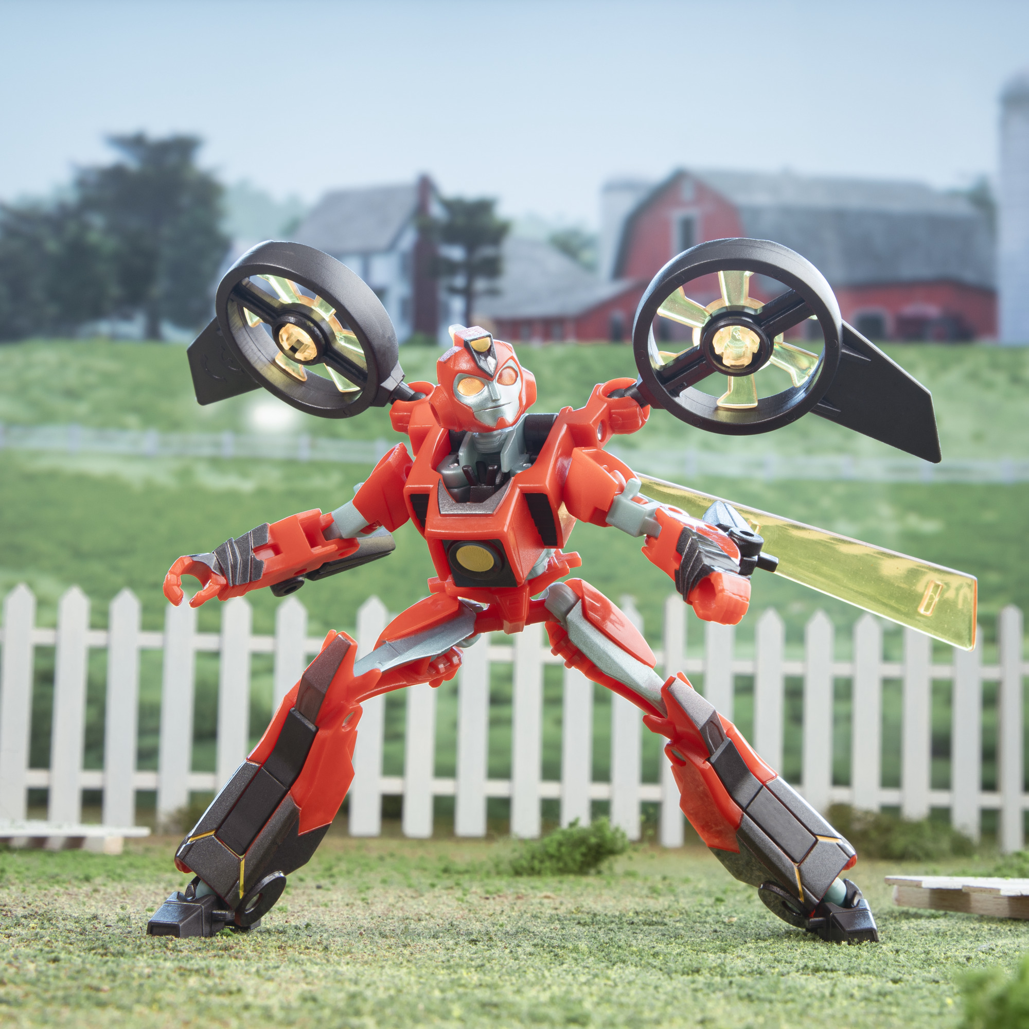 Un giocattolo robot rosso è in piedi sull'erba di fronte a una staccionata bianca, una fattoria sullo sfondo e due oggetti neri.
