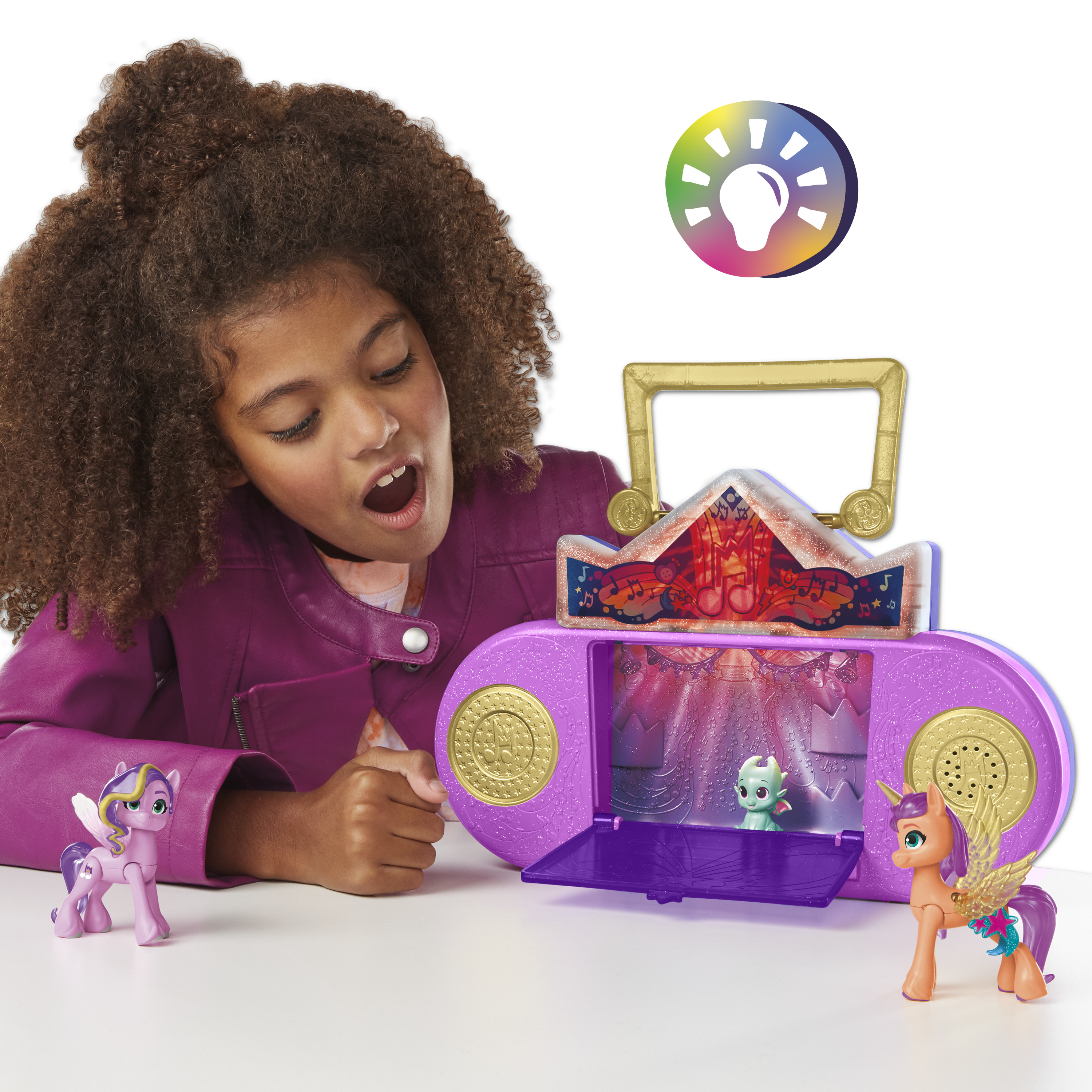 Dziewczyna patrzy na zabawkowe podium z figurkami. Scena jest otwarta, z małą figurką w środku. Dwie figurki kucyków stoją.