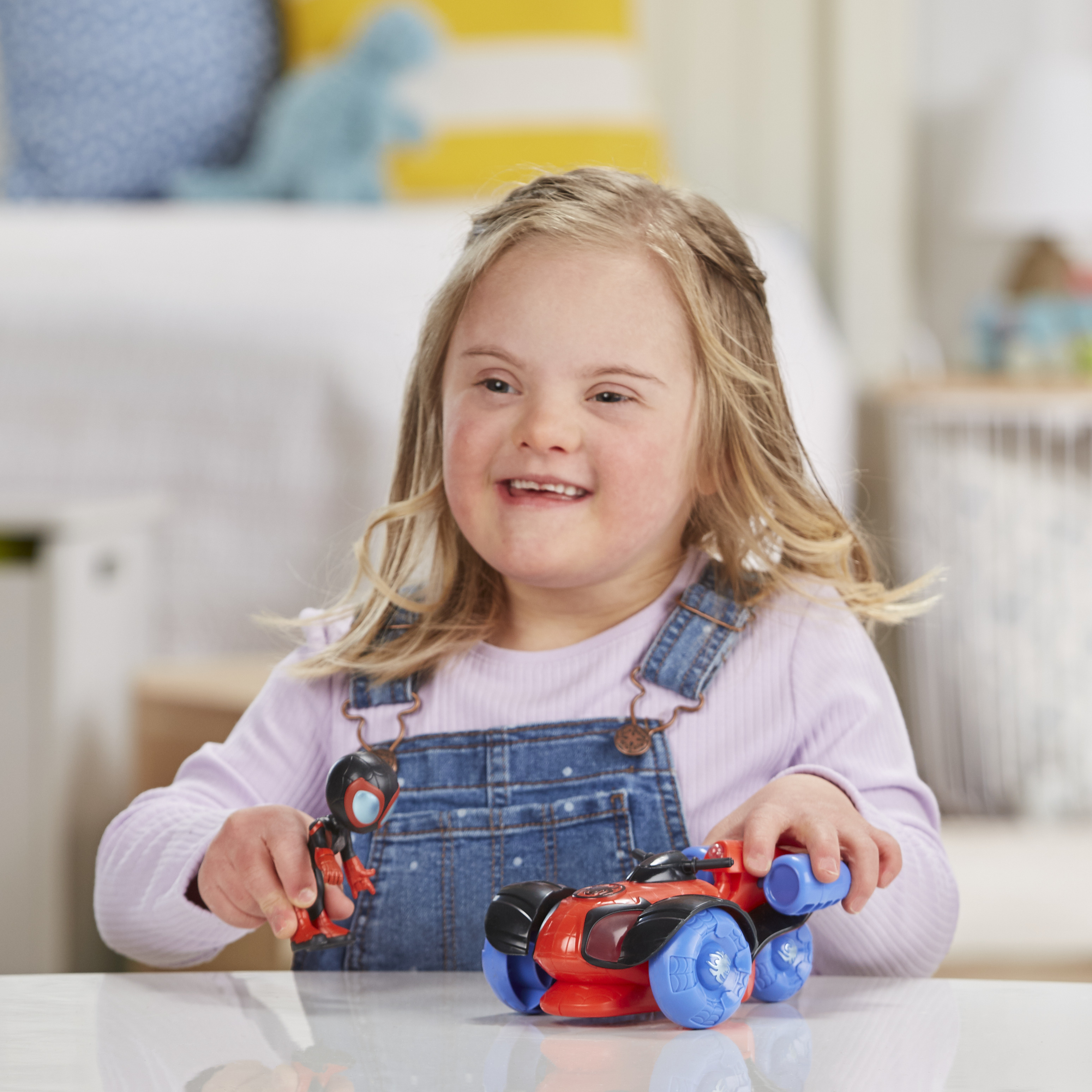 Uśmiechnięta dziewczynka z zespołem Downa trzyma zabawki. Jedna to figurka Spider-Mana, druga czerwono-niebieski pojazd.