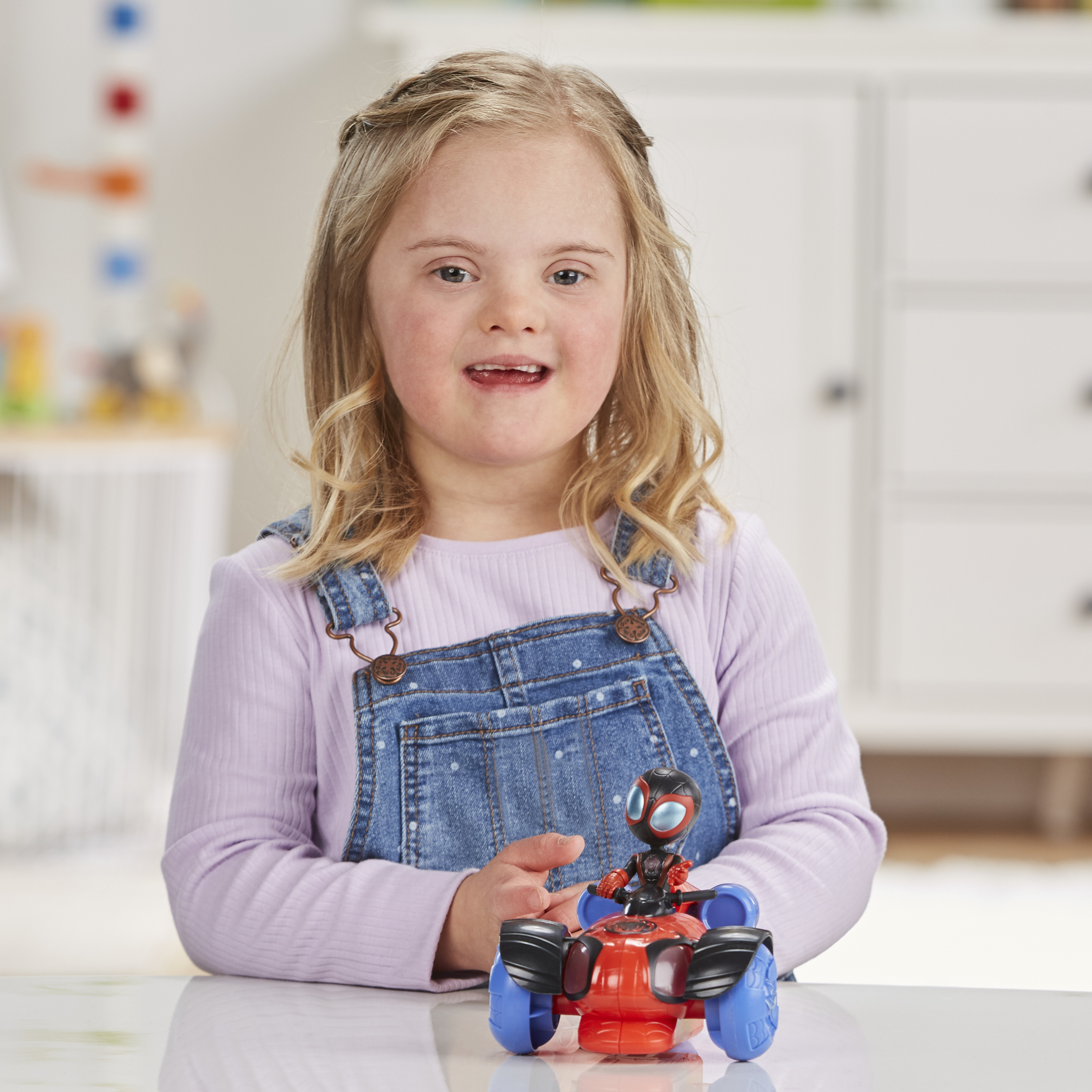 Dziewczyna z blond włosami uśmiecha się. Ma na sobie dżinsowe ogrodniczki i trzyma czerwoną zabawkę.