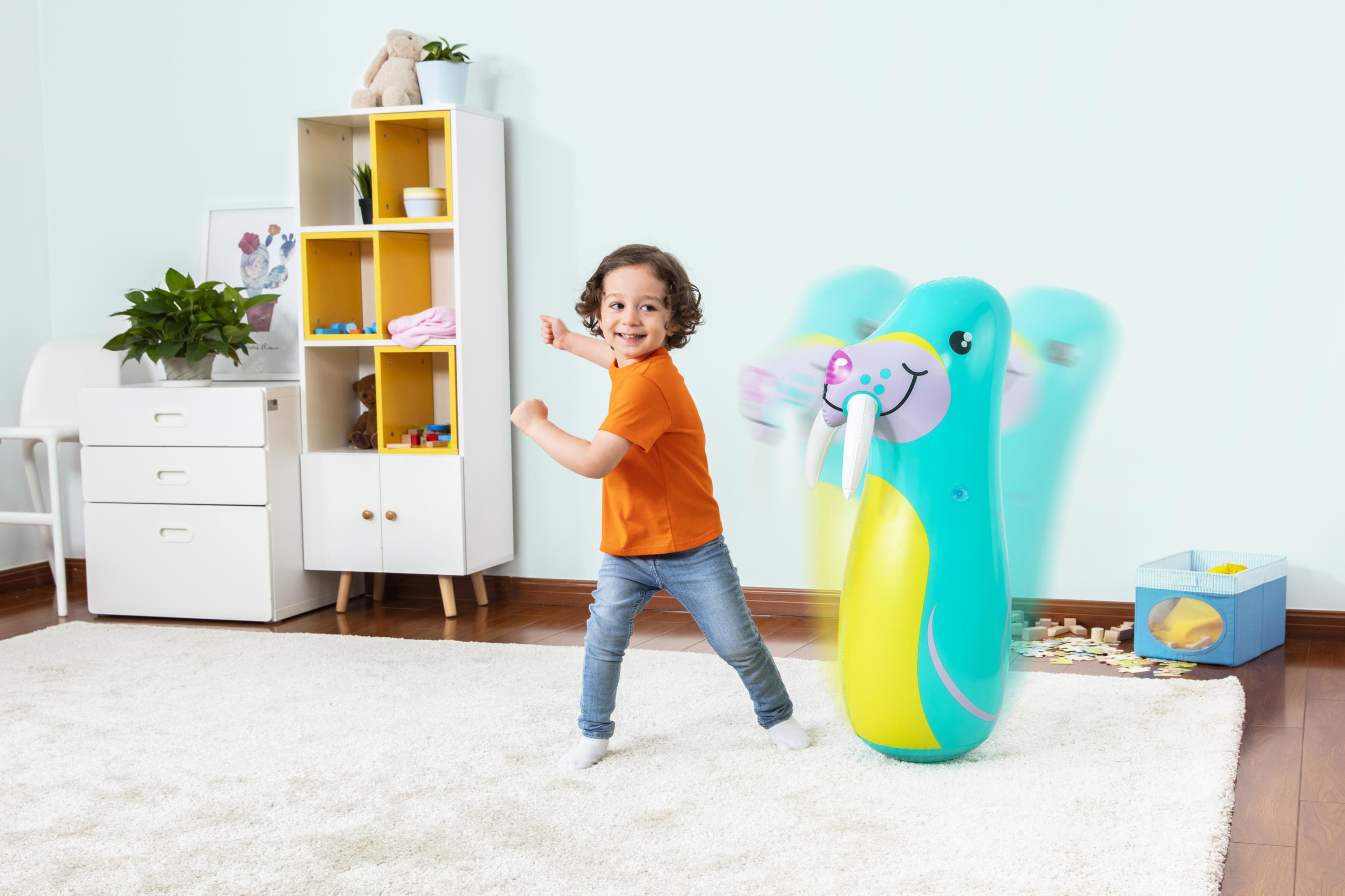 Dziecko w pomarańczowej koszulce bawi się nadmuchiwaną zabawką w kształcie foki. W tle biała szafka.