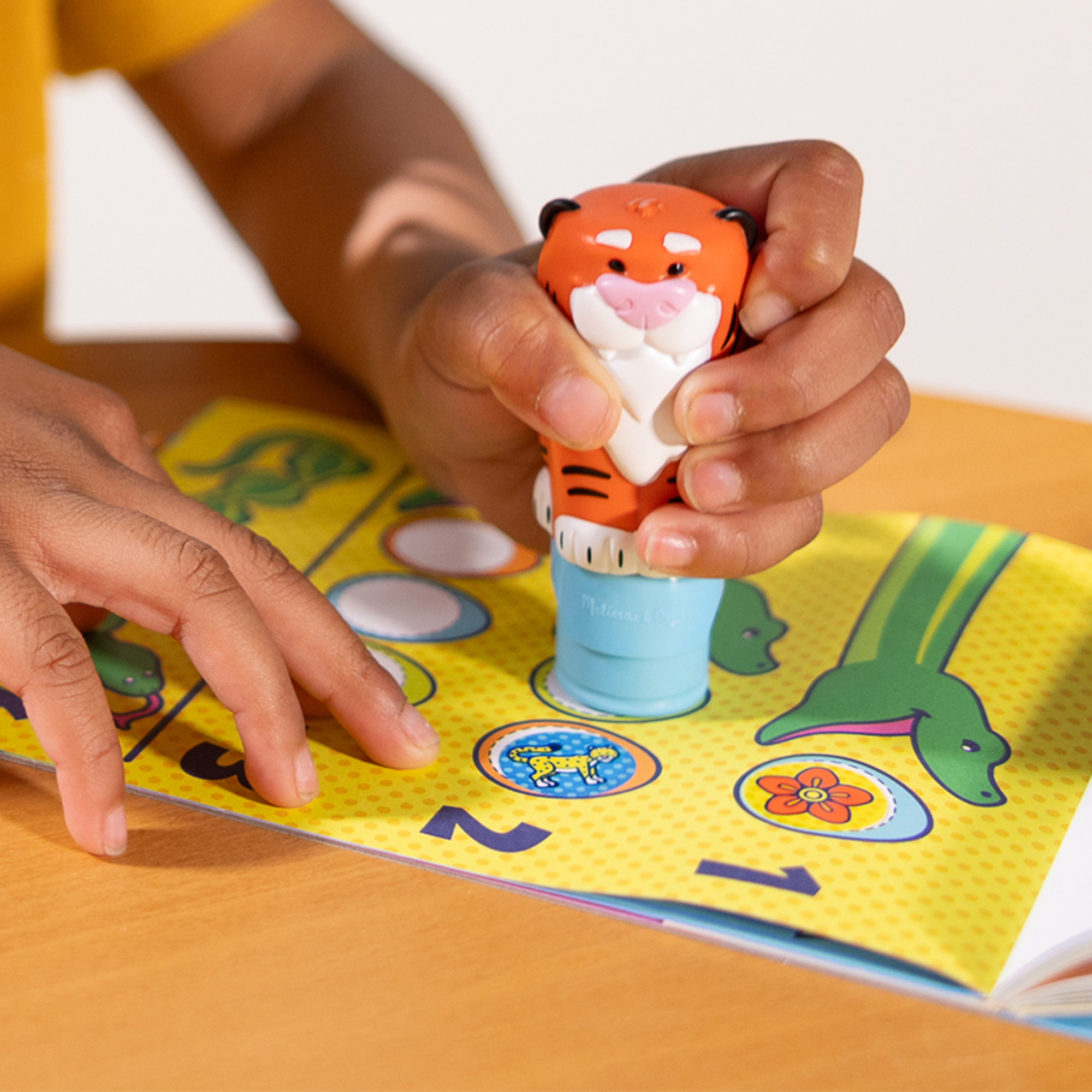 Kinderhände halten einen Tigerstempel über einem farbenfrohen Spielbrett. Das Brett hat Tierillustrationen.