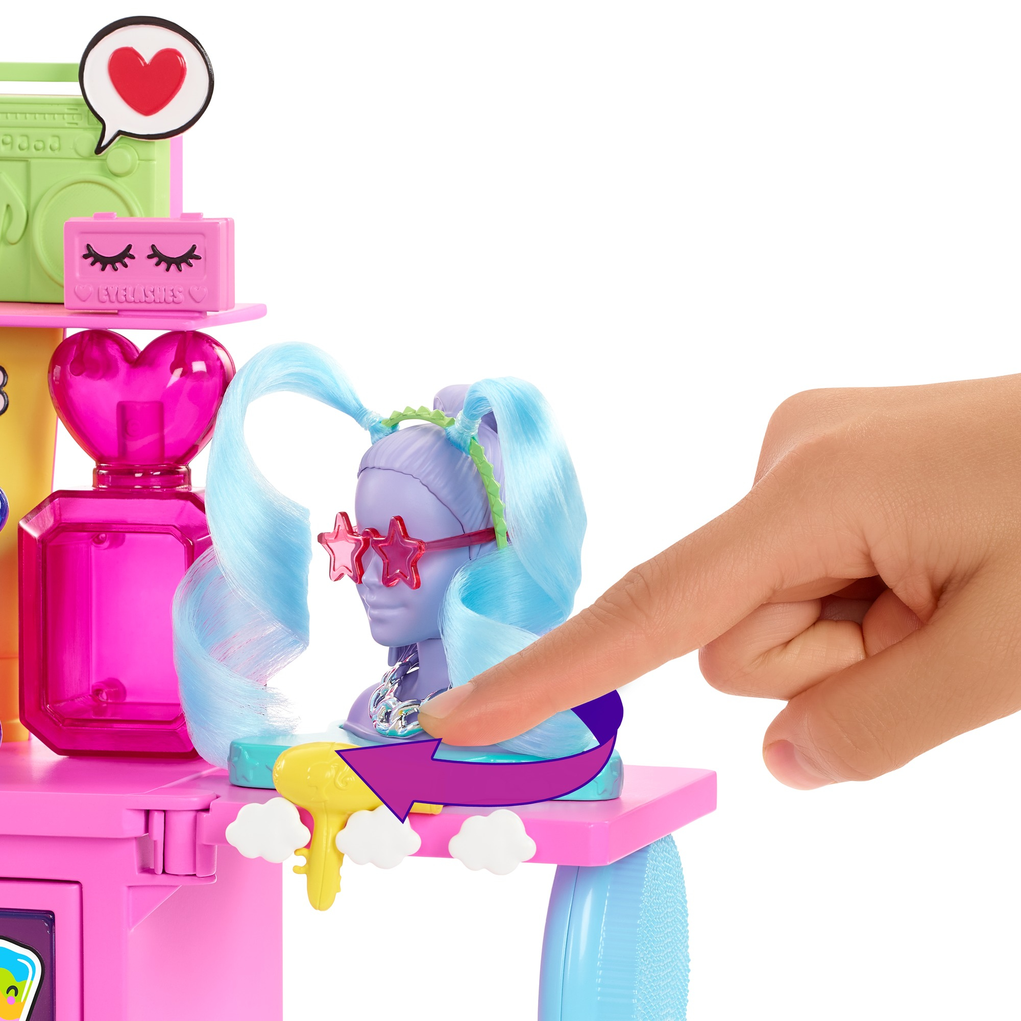 Eine Hand zeigt auf einen Puppenkopf mit blauem Haar und Sternbrille. Im Hintergrund sind rosa und grüne Spielzeugstrukturen zu sehen.