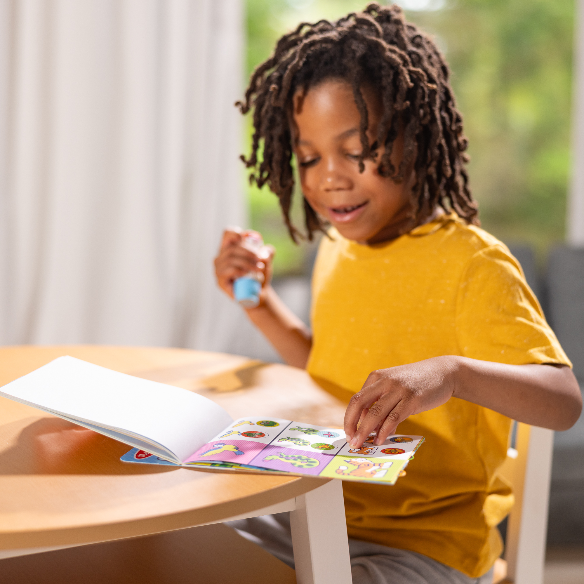 Ein Junge mit Dreadlocks betrachtet ein Buch mit Aufklebern. Er hält ein kleines Spielzeug, während er an einem Tisch sitzt.