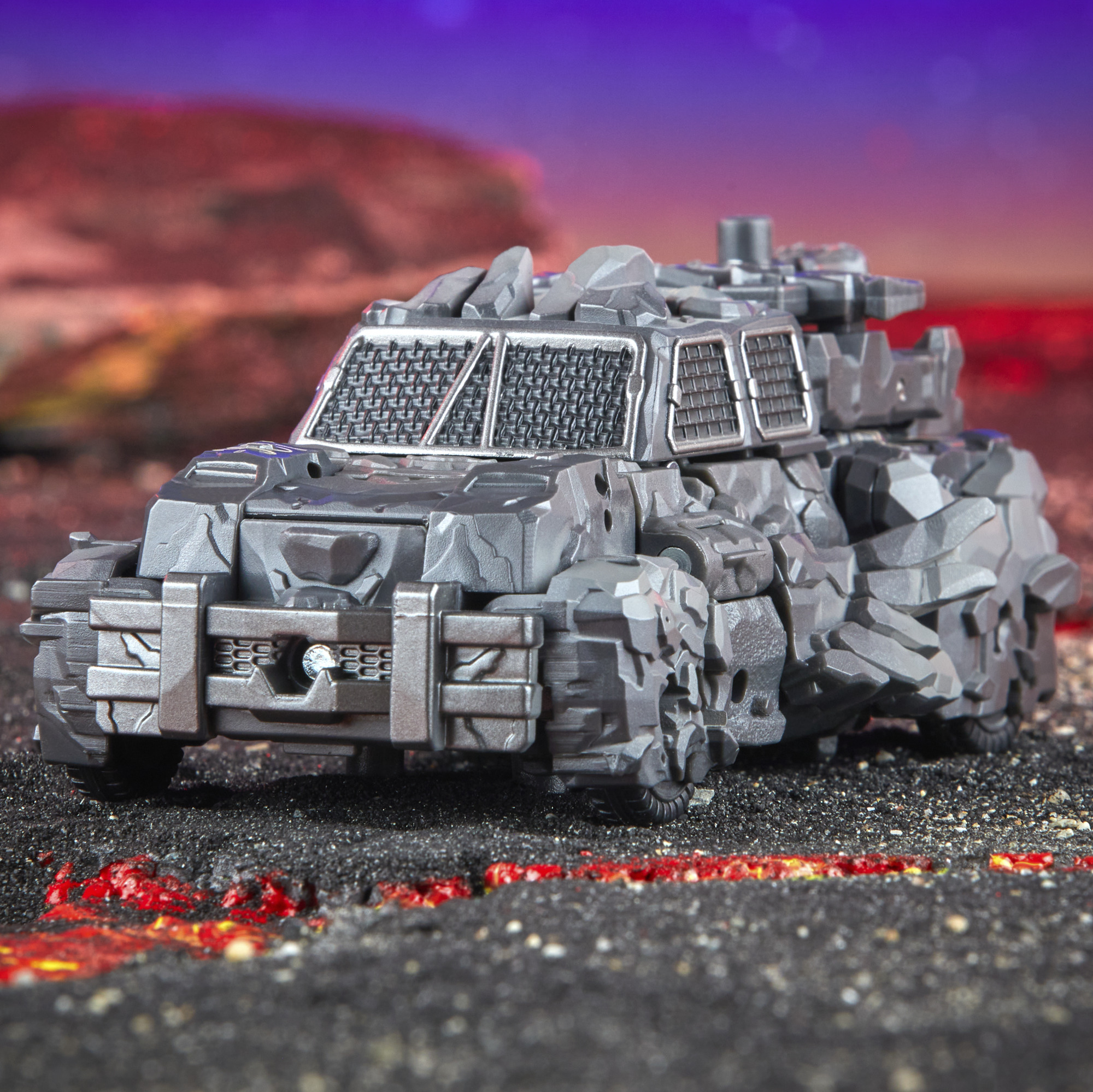 Szara zabawka w postaci opancerzonego pojazdu z kamienistymi fakturami stoi na ciemnej powierzchni z czerwonymi smugami, fioletowym i czerwonym tłem.