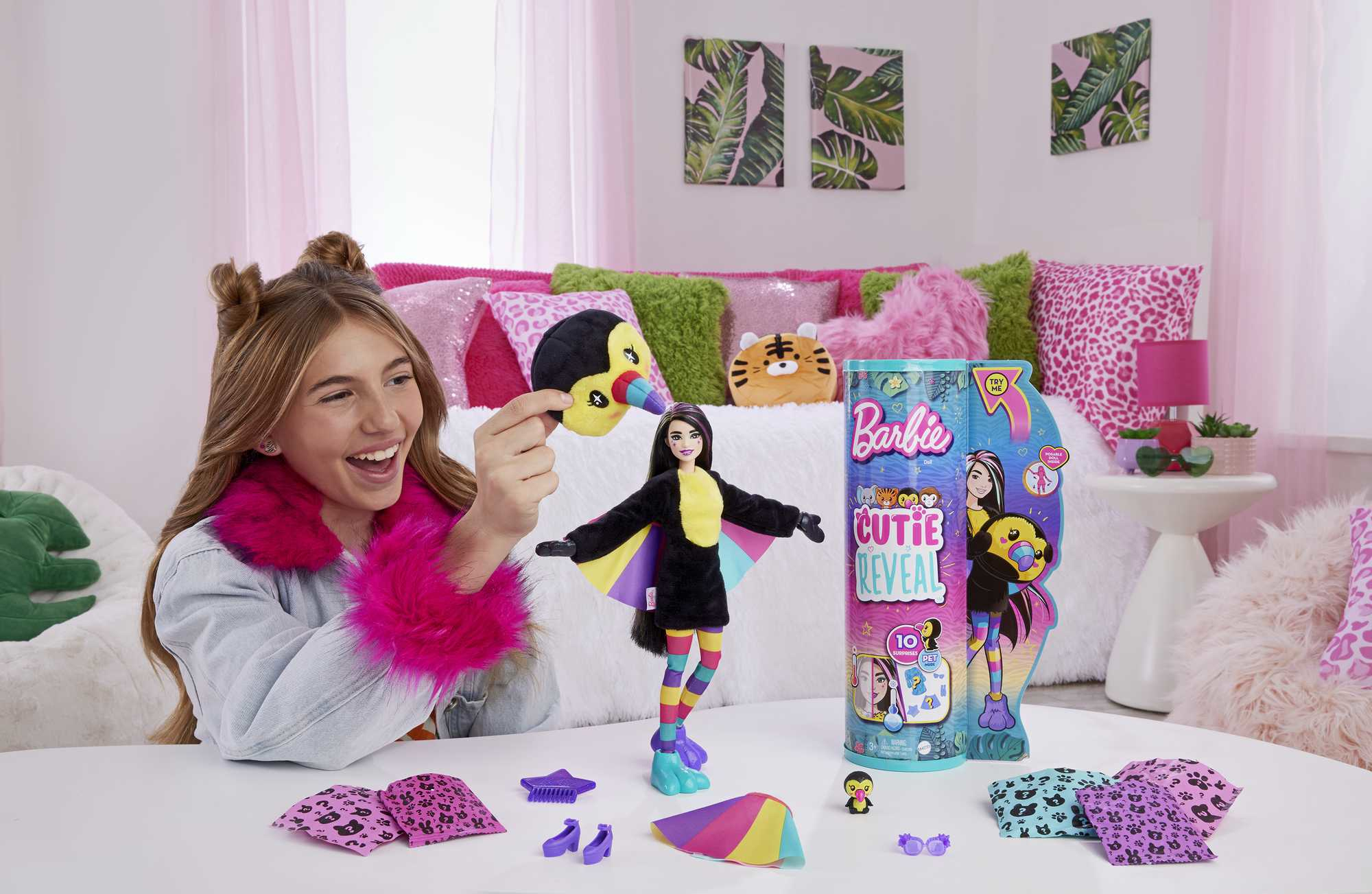 Ein Mädchen zeigt eine Spielzeugpuppe in einem Pinguinkostüm. Verschiedenes Spielzeugzubehör liegt auf dem Tisch, und eine Barbie-Box ist im Hintergrund.