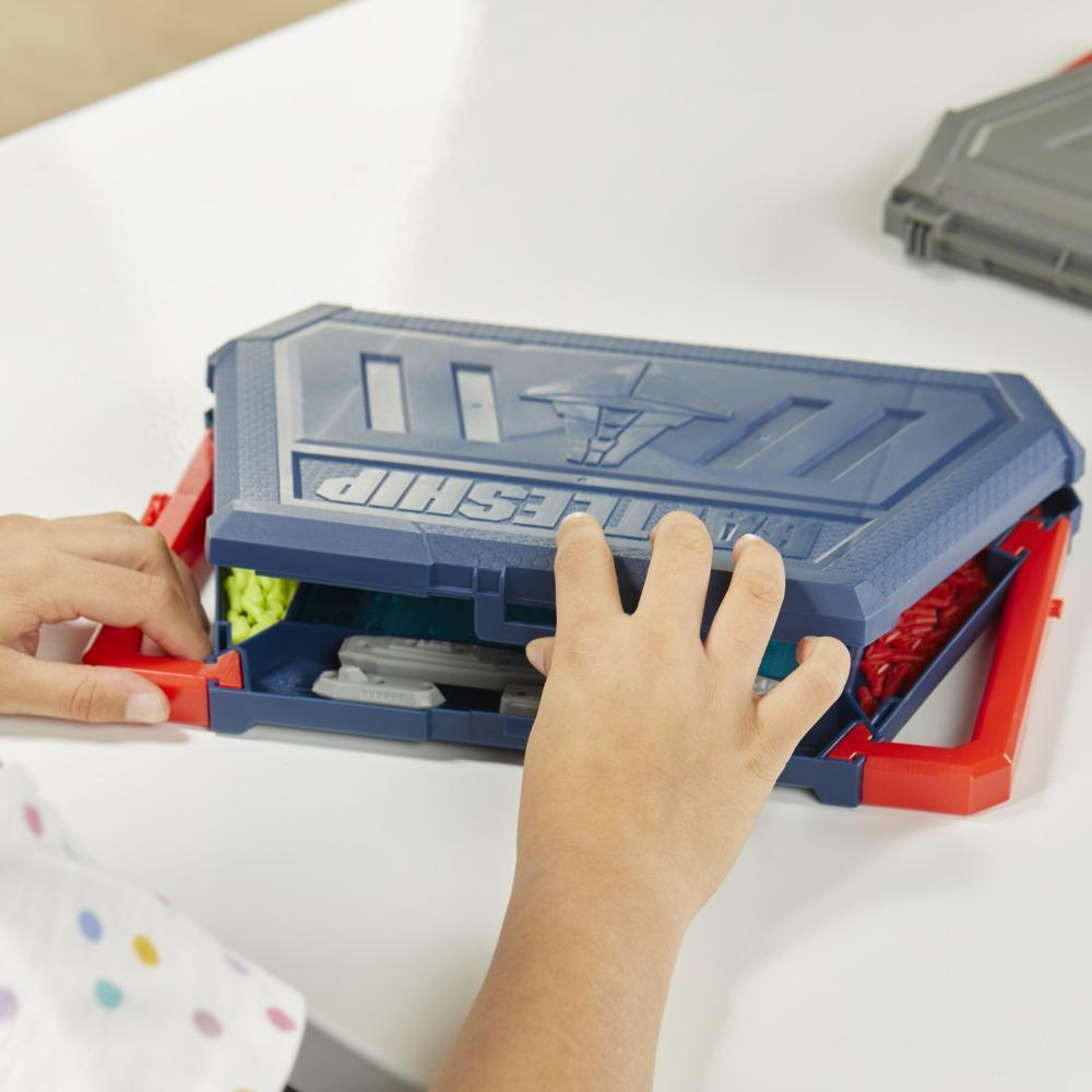 Las manos de un niño abren un estuche de juego de Batalla Naval. El estuche es azul y rojo, con el nombre. El fondo es una mesa blanca.