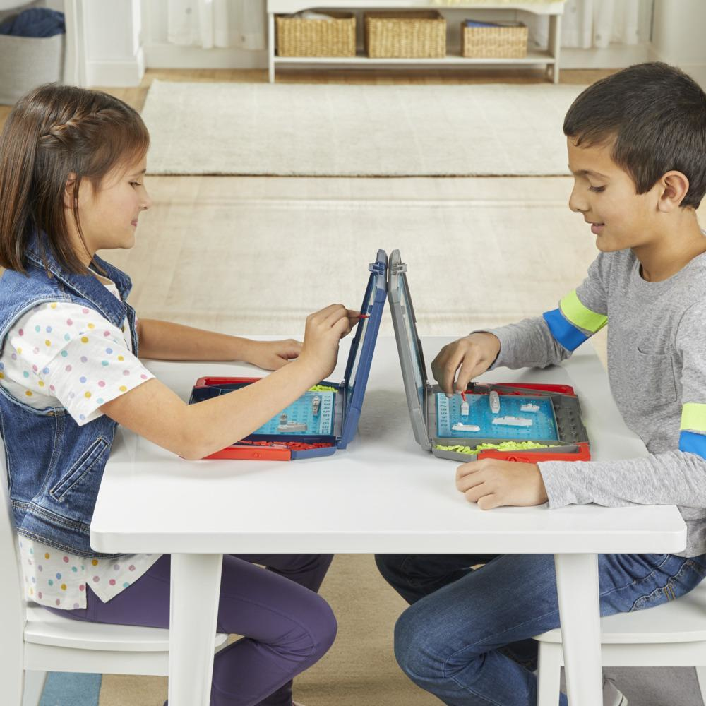 Dos niños juegan a un juego de mesa, sentados en una mesa blanca. Ambos se concentran en sus tableros, cada uno con barcos. Un juego de batalla naval.