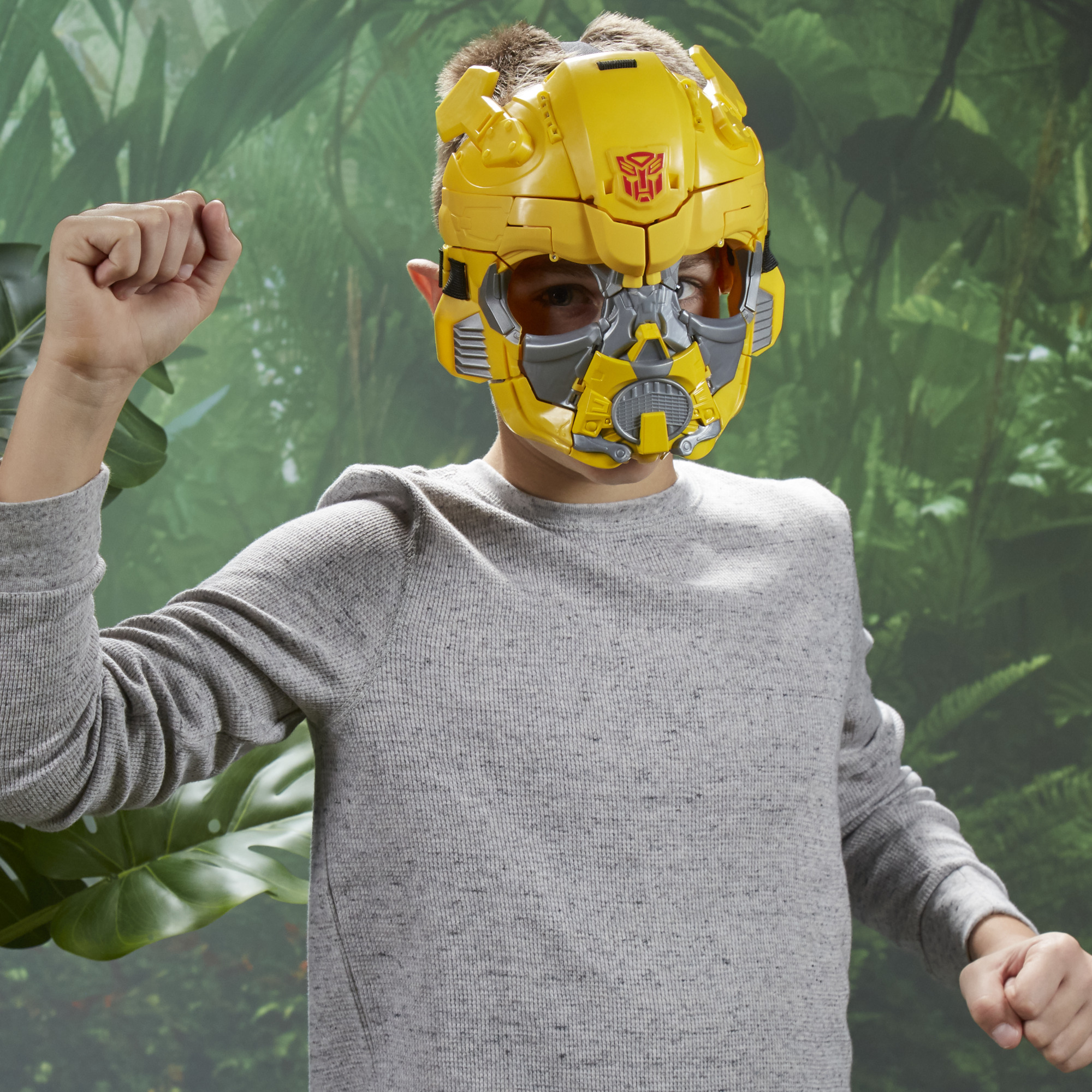 Un niño con una máscara de Bumblebee levanta el puño. El fondo es un papel pintado con temática de jungla. El niño lleva un suéter gris.