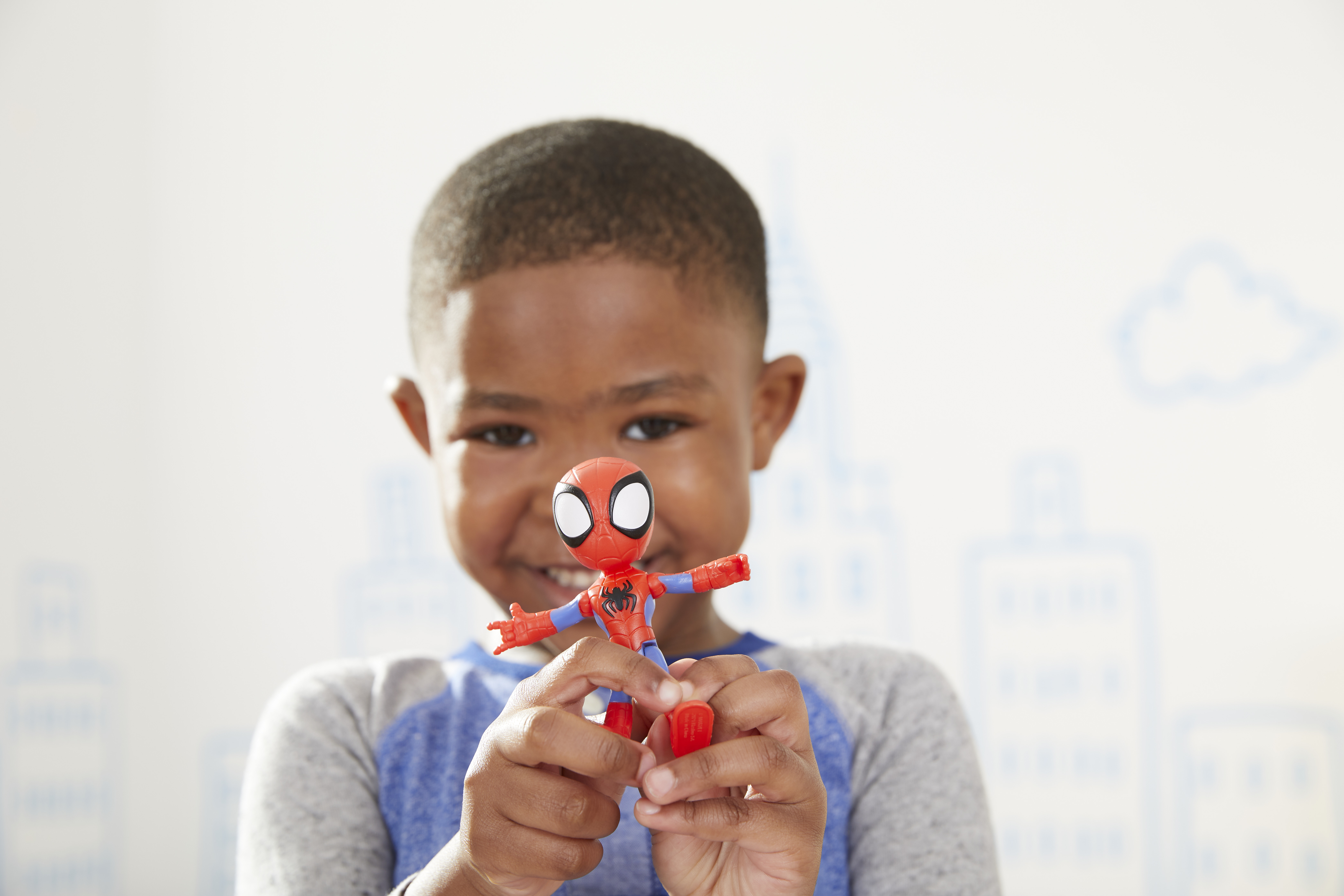 Un niño sostiene un juguete de Spider-Man. El fondo tiene un contorno de ciudad y una nube. El juguete es rojo, azul y negro.