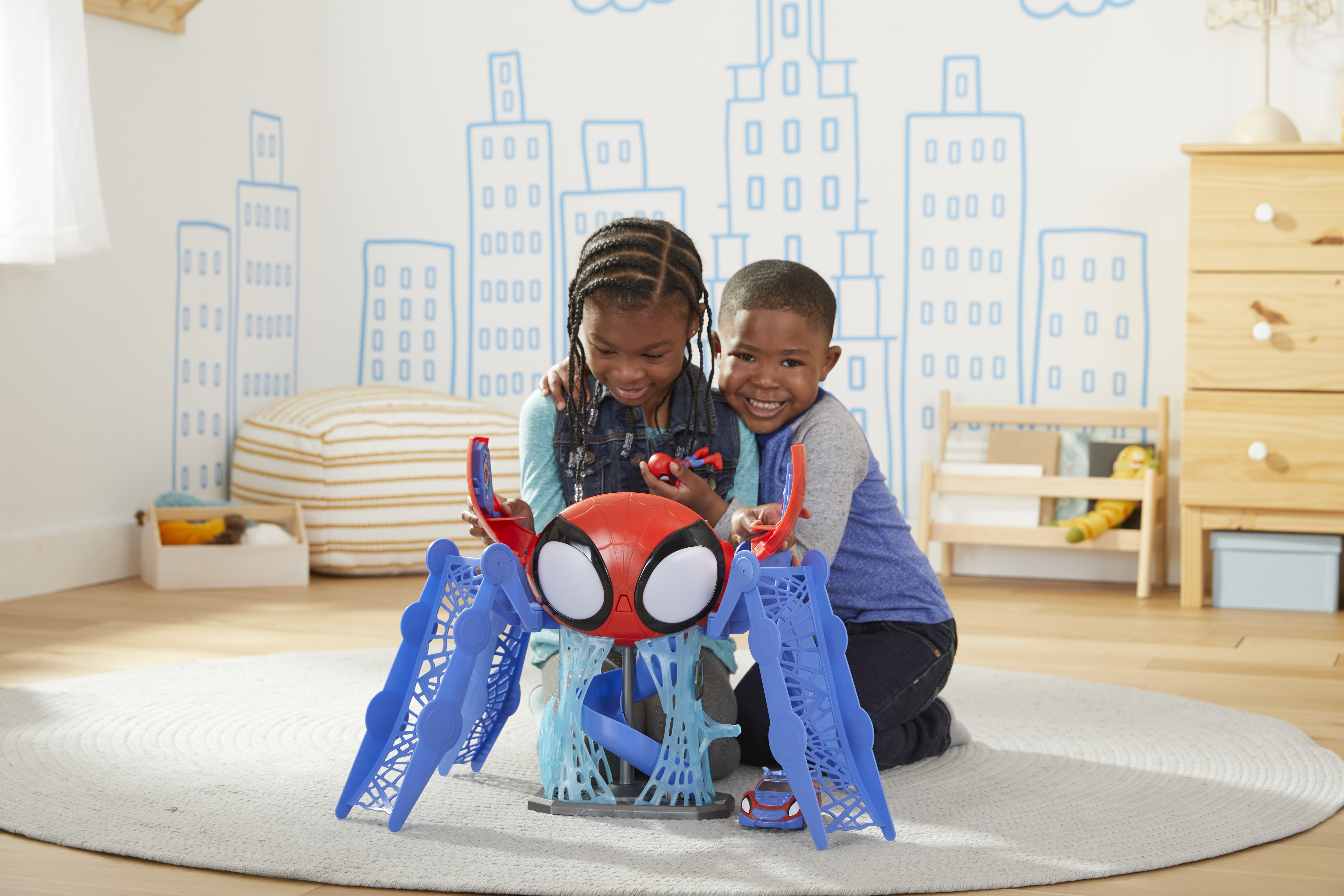 Dos niños juegan con un juguete de Spider-Man. El juguete es rojo y azul. El fondo es una habitación con dibujos de la ciudad.