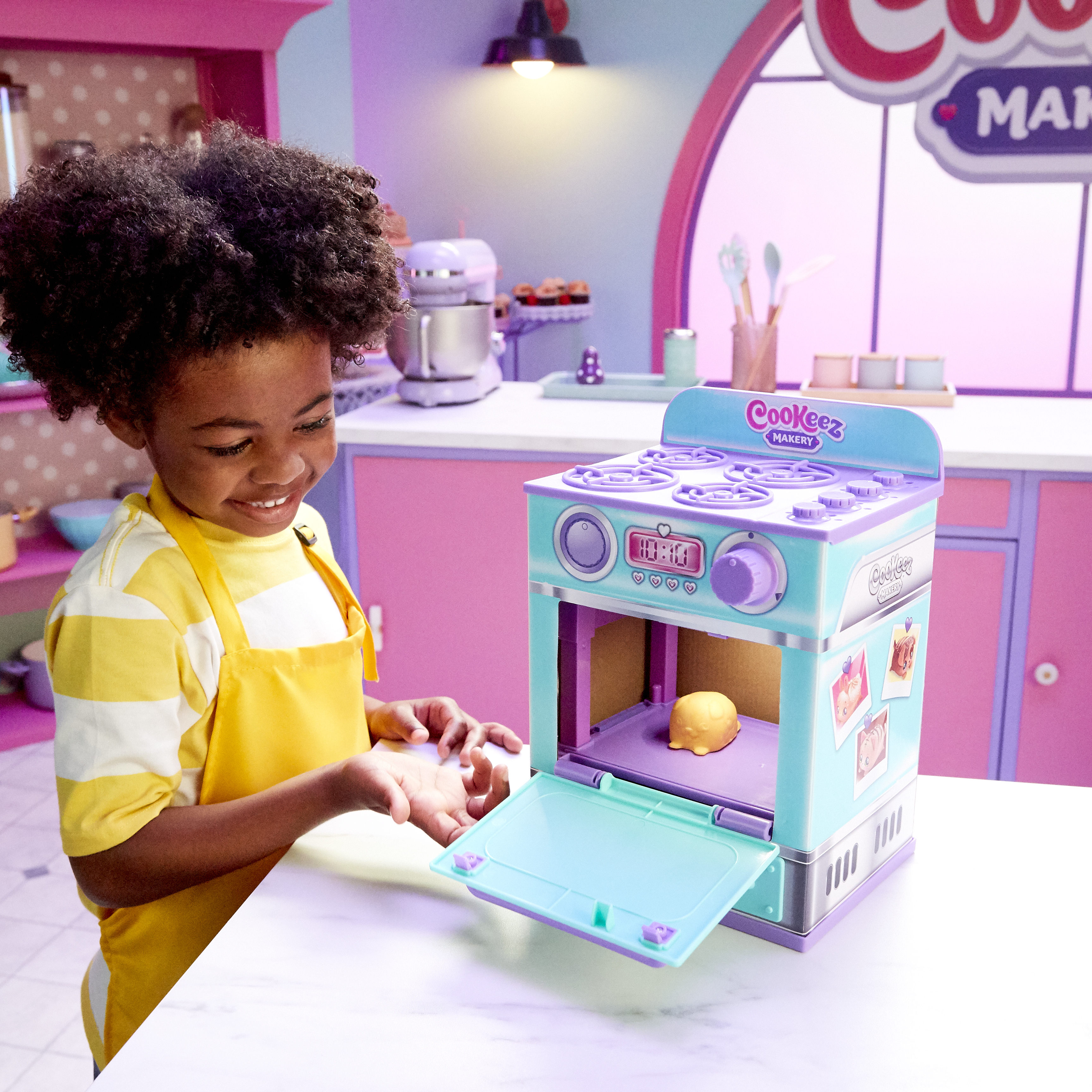 Dziecko w żółtym fartuchu uśmiecha się do zabawkowego piekarnika. W środku żółte ciasteczko. Scena to zabawka kuchnia.