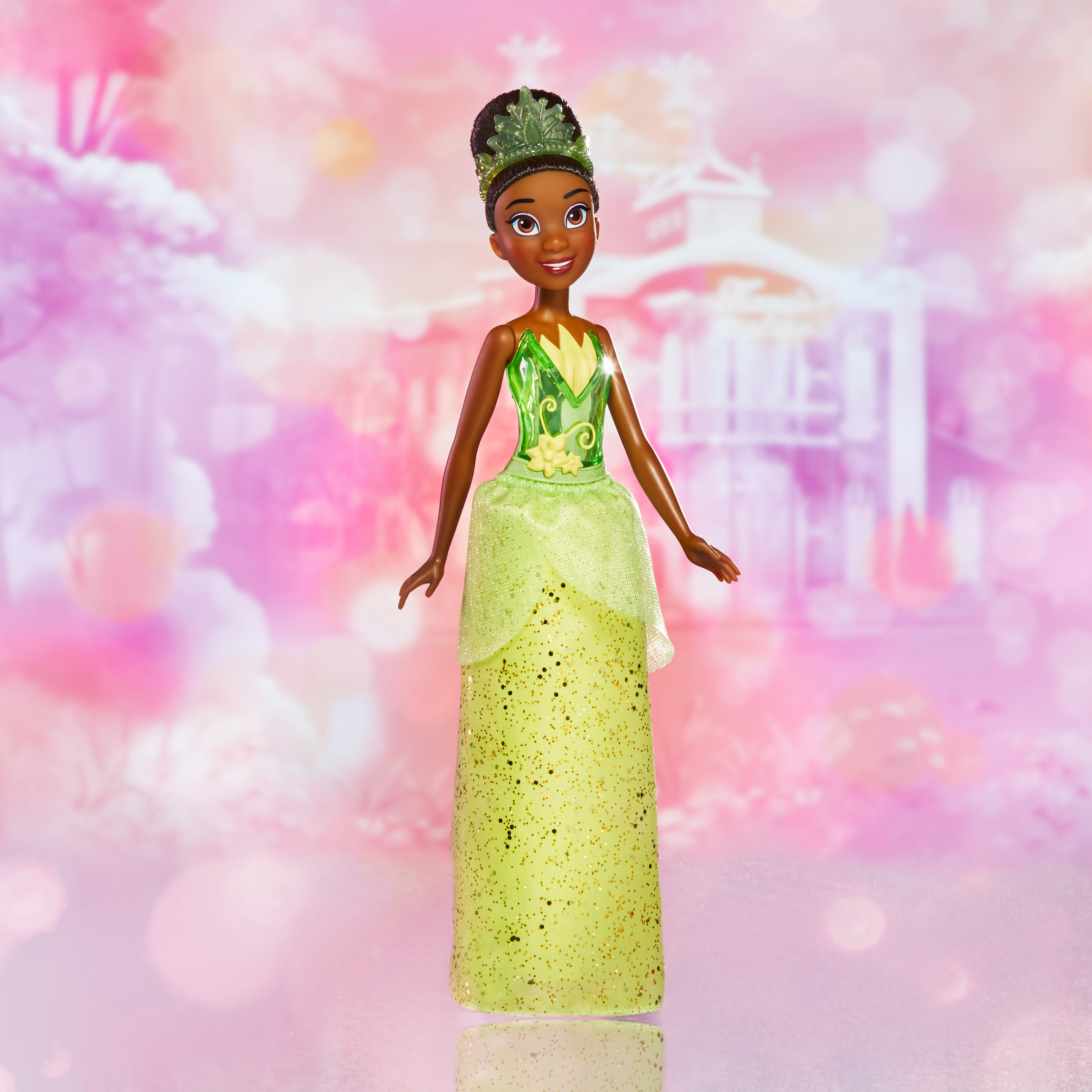 Una bambola di Tiana è in piedi. Indossa un vestito verde e una corona. Lo sfondo è rosa con un castello.