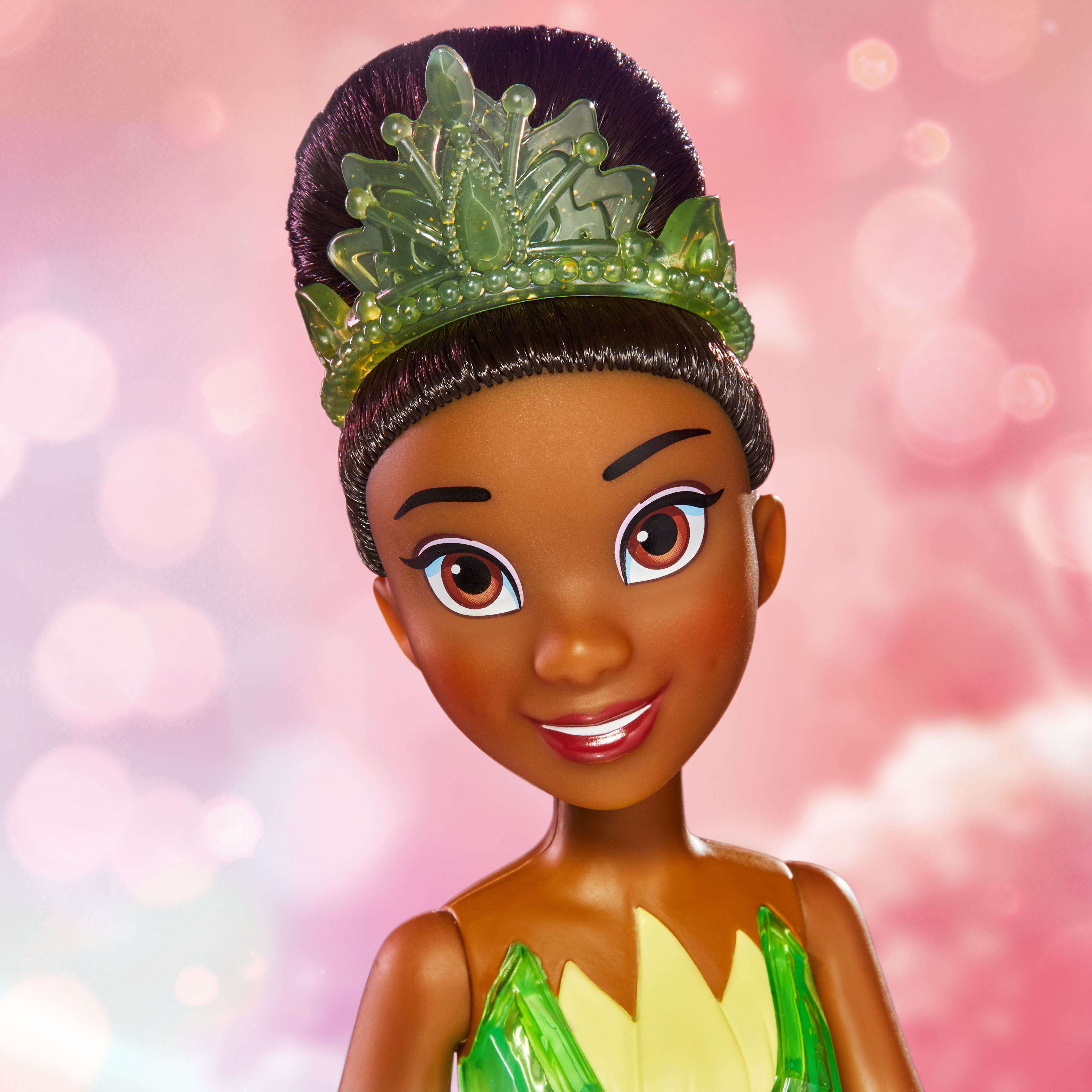 Bambola di Tiana con capelli scuri e tiara verde. Indossa un vestito verde e giallo. Sfondo rosa bokeh.
